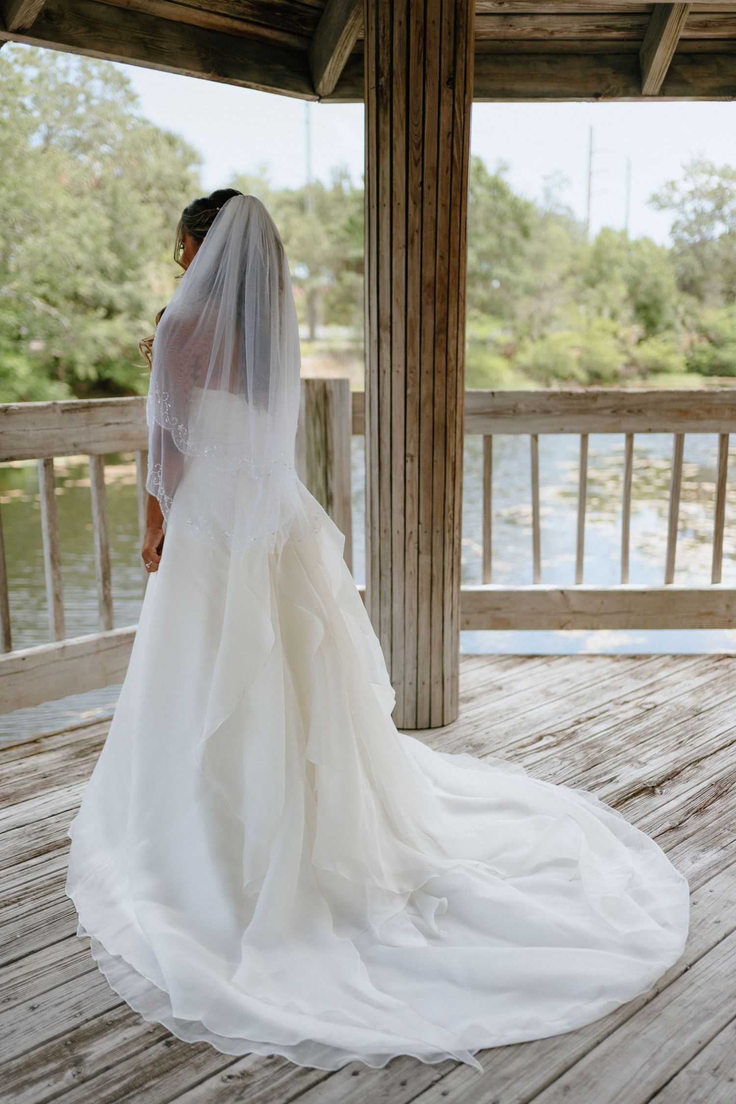 Bride in a white ballgown with long veil stands with her back to camera on a deck.
