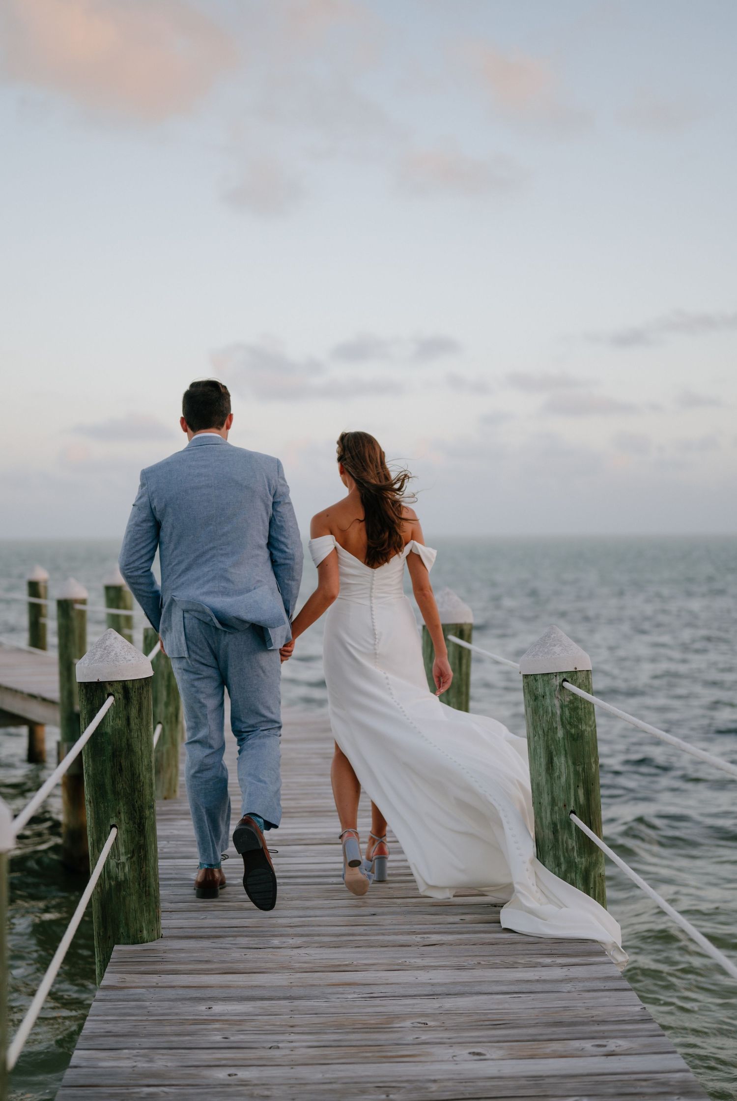 Bride and groom walking hand in hand toward the ocean on a wooden pier at a waterfront wedding.