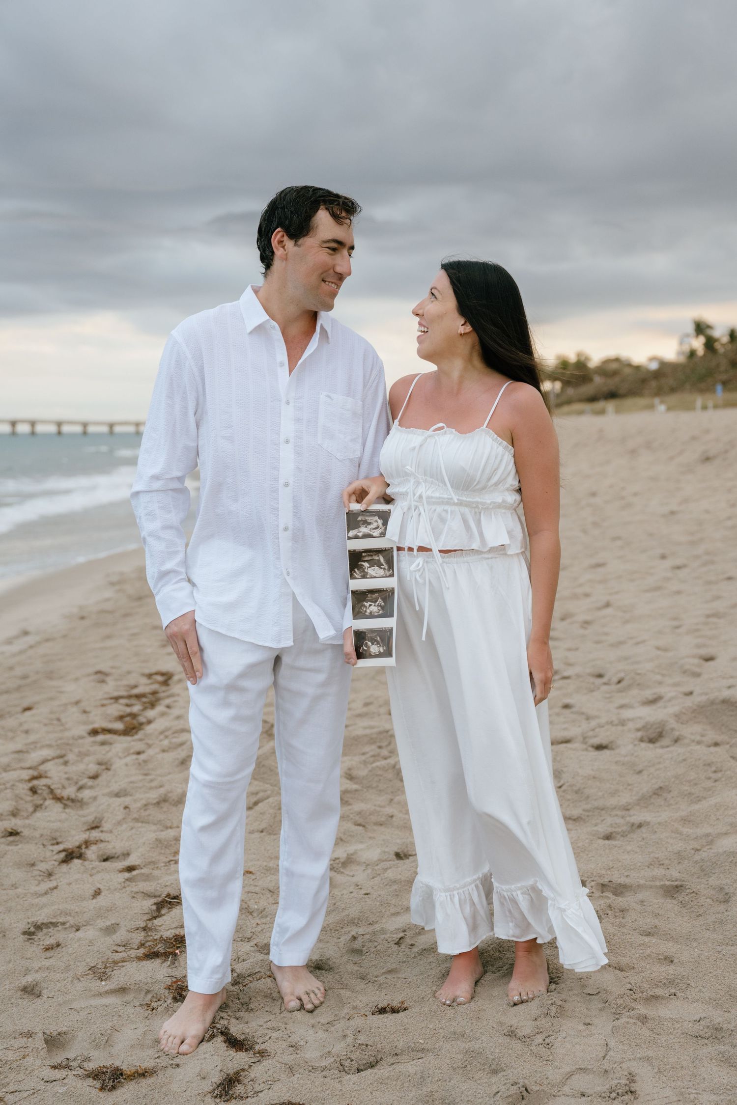 Couple in matching white outfits shares a smile while holding ultrasound photos on a cloudy beach.