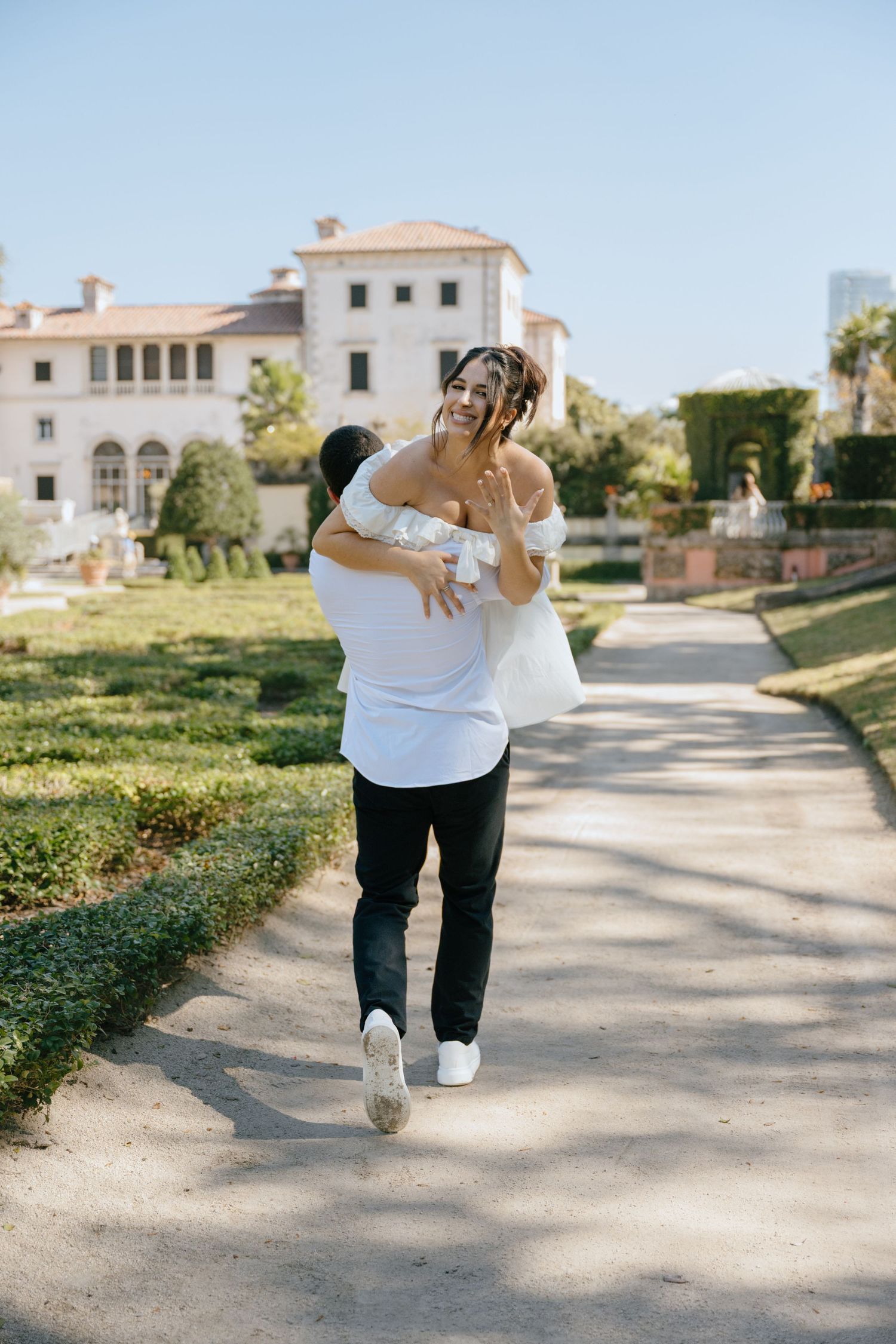 Groom carries laughing bride in white dress along a garden path near an elegant estate.