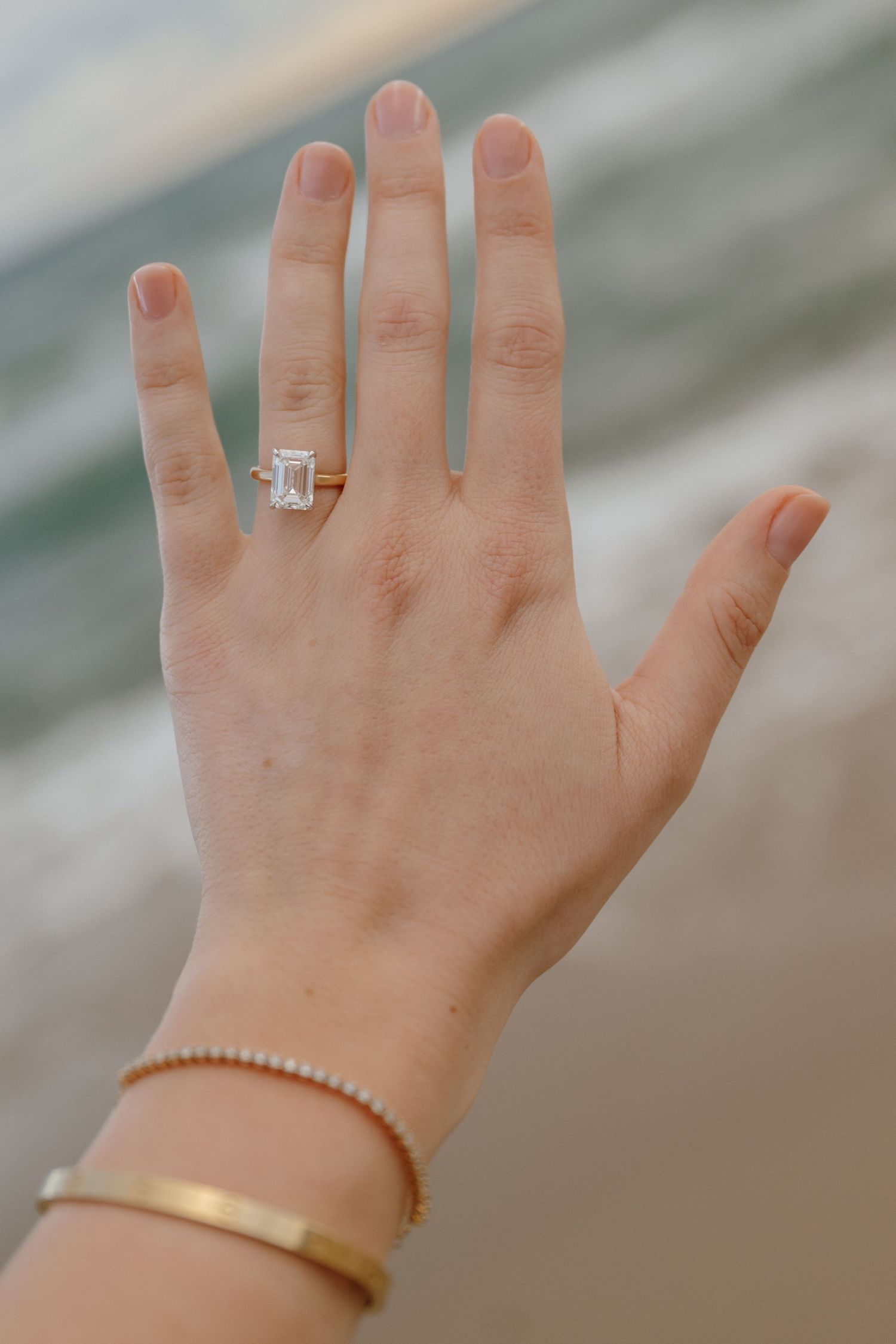 Close-up of hand displaying emerald cut diamond engagement ring with gold bracelet at beach.