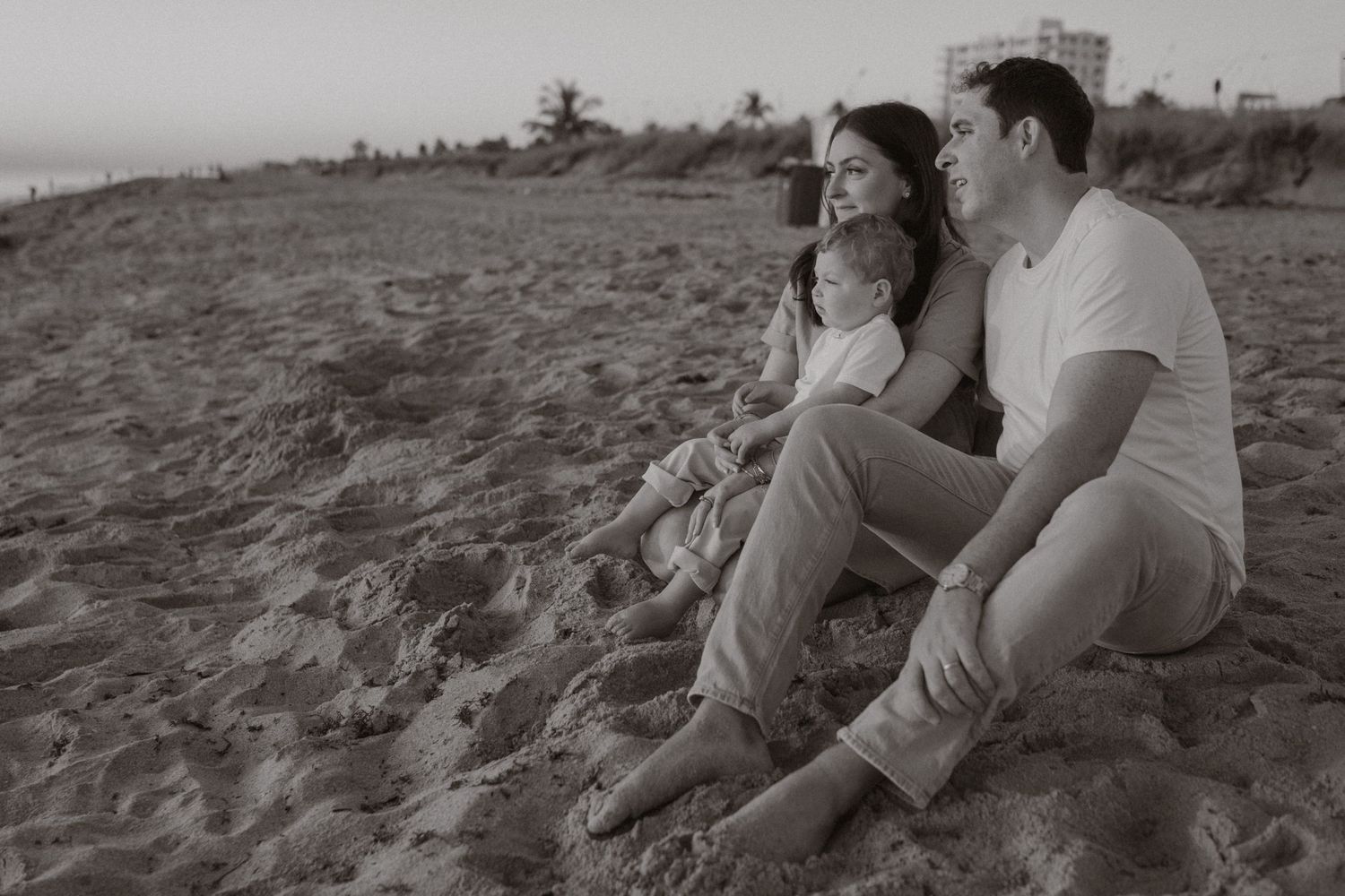 Black and white photo of a couple sitting on the beach with their young child at dusk.