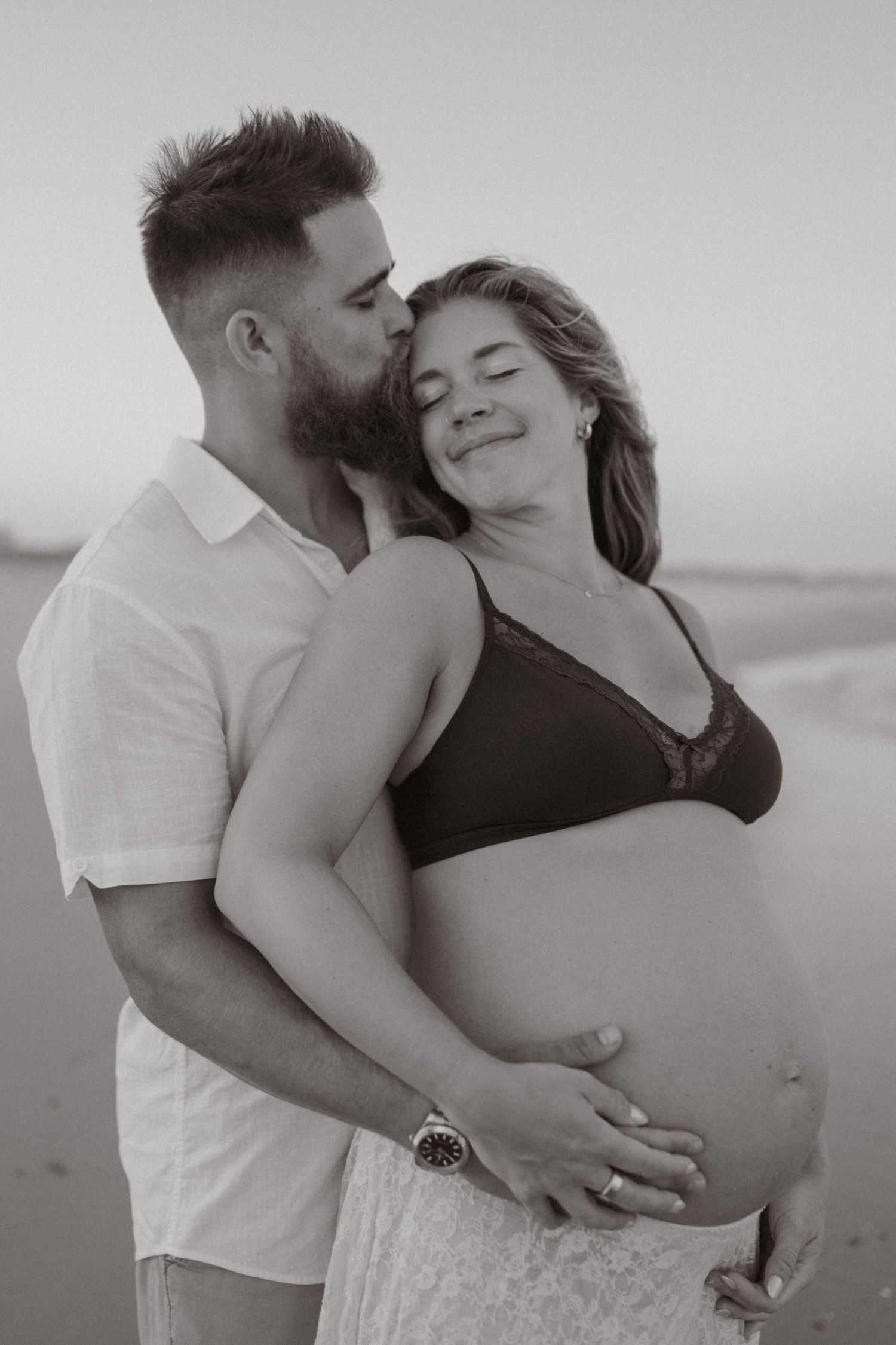 Black and white photo of bearded man kissing pregnant woman's cheek on beach.