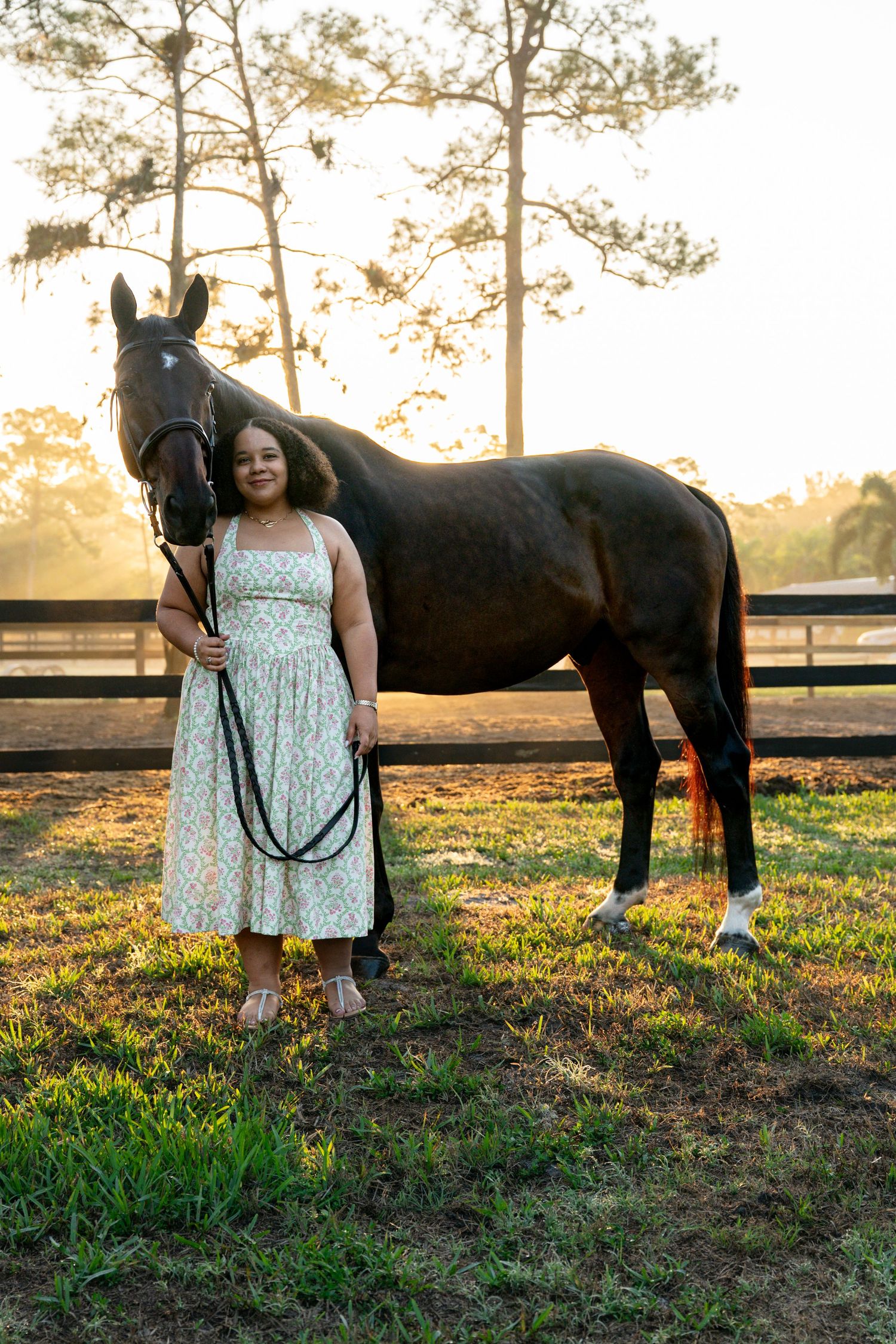 Woman in floral dress stands with a dark bay horse at golden hour sunset in a paddock.