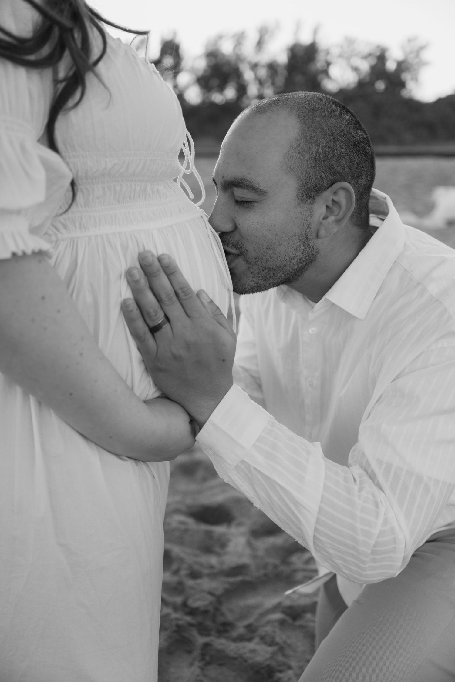 Black and white image of man kneeling to kiss pregnant woman's belly on sandy beach.