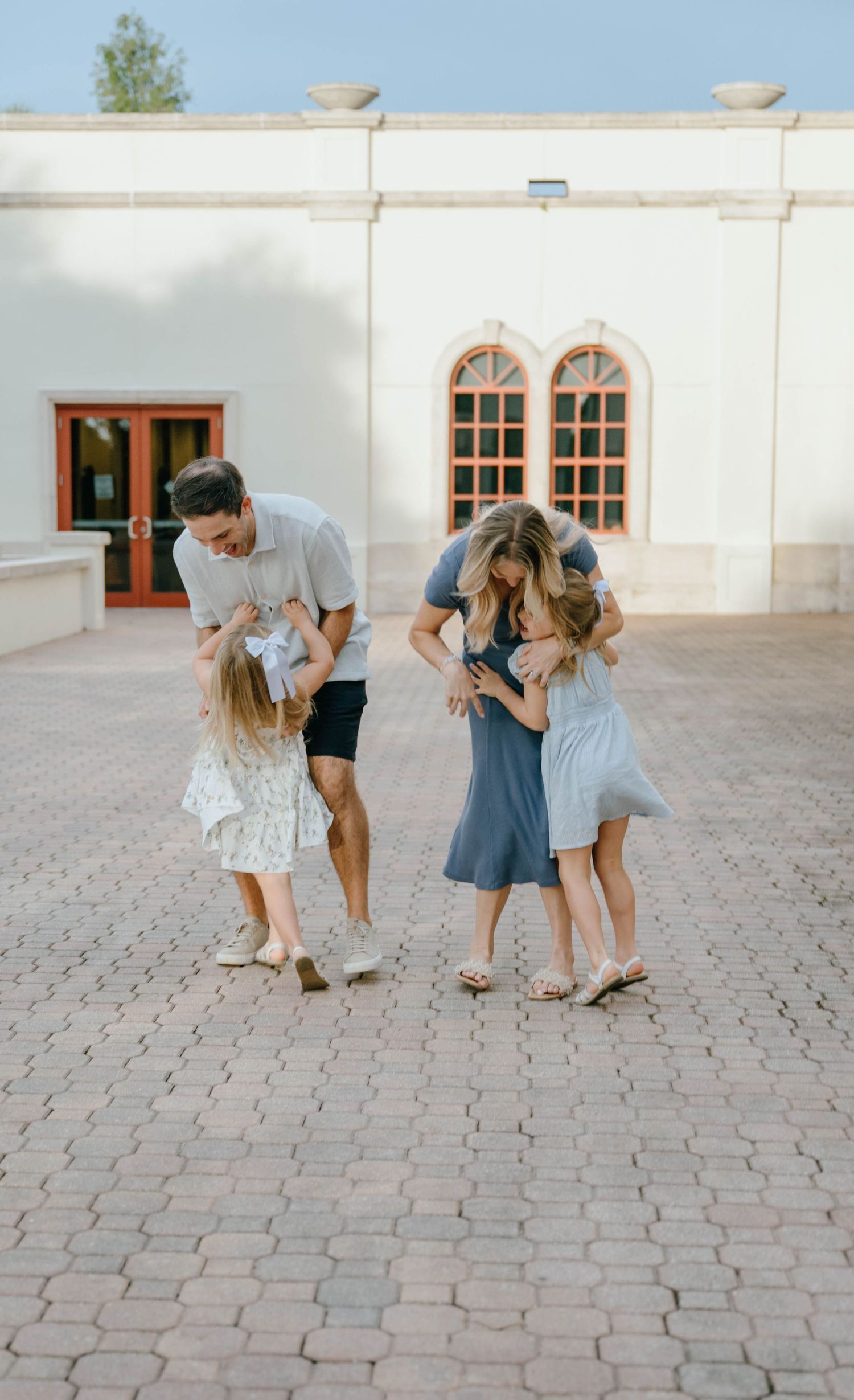 A joyful family of four embraces and plays together on a brick courtyard outside a building.