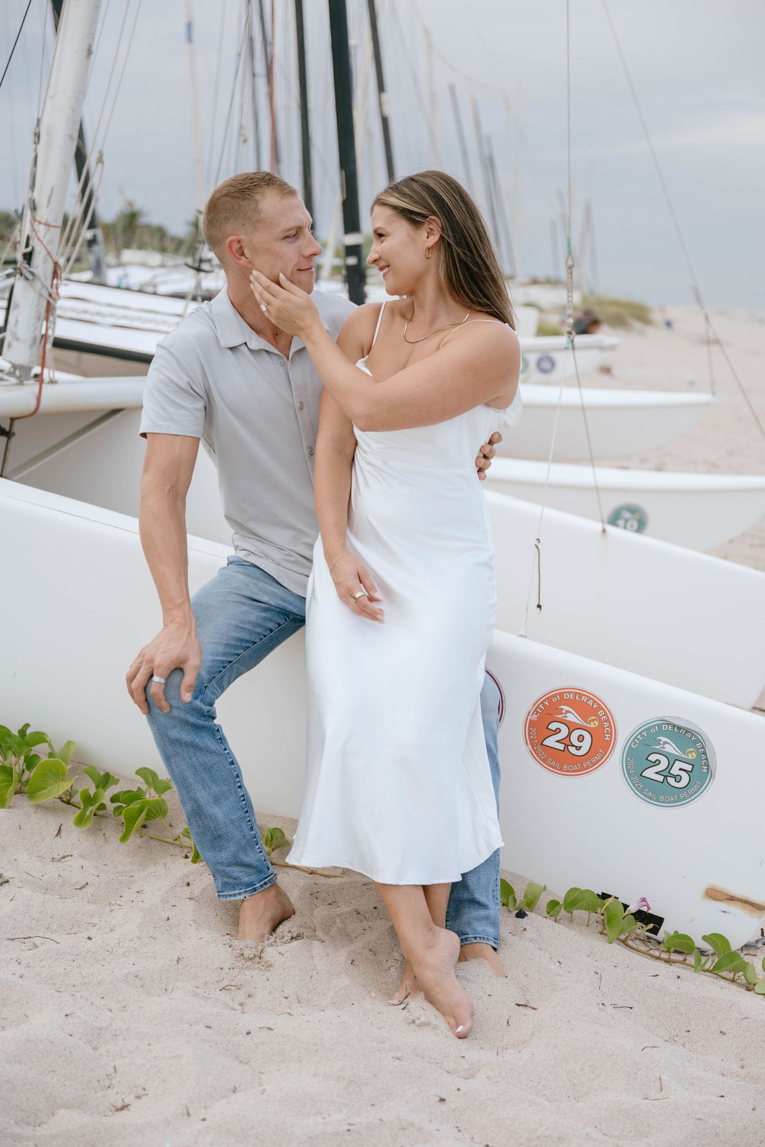 Happy couple sharing a tender moment beside numbered sailboats on a sandy beach at dusk.