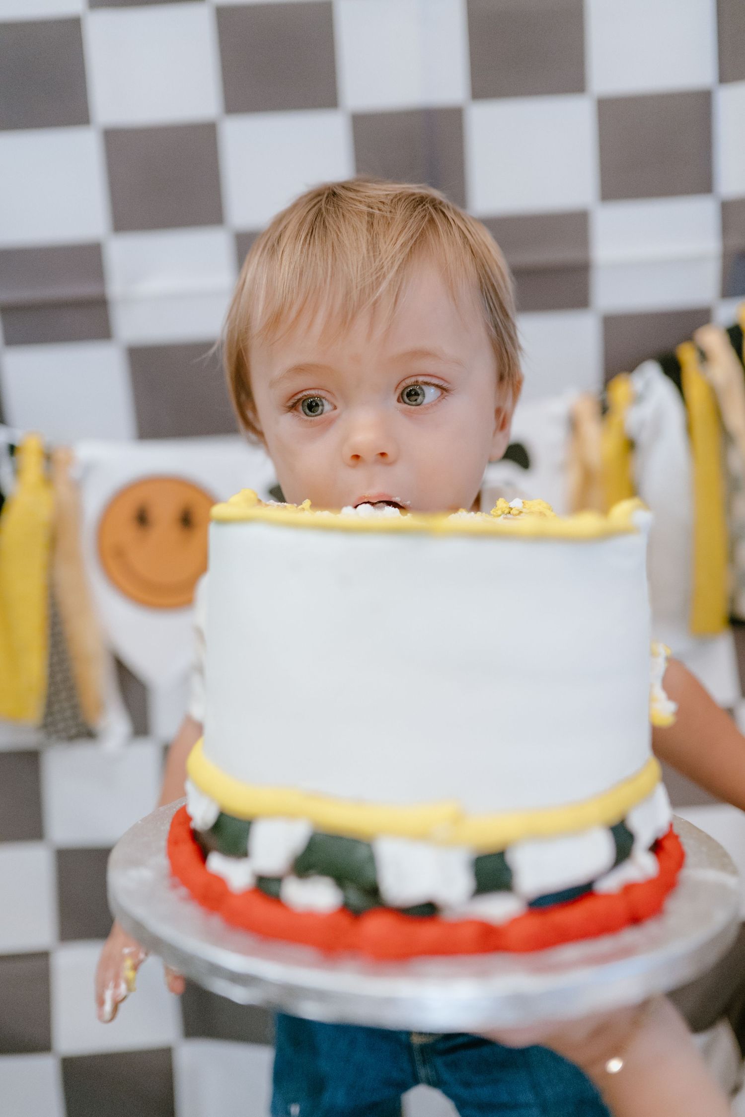 Wide-eyed toddler peeks over a white and yellow first birthday smash cake held up for him.
