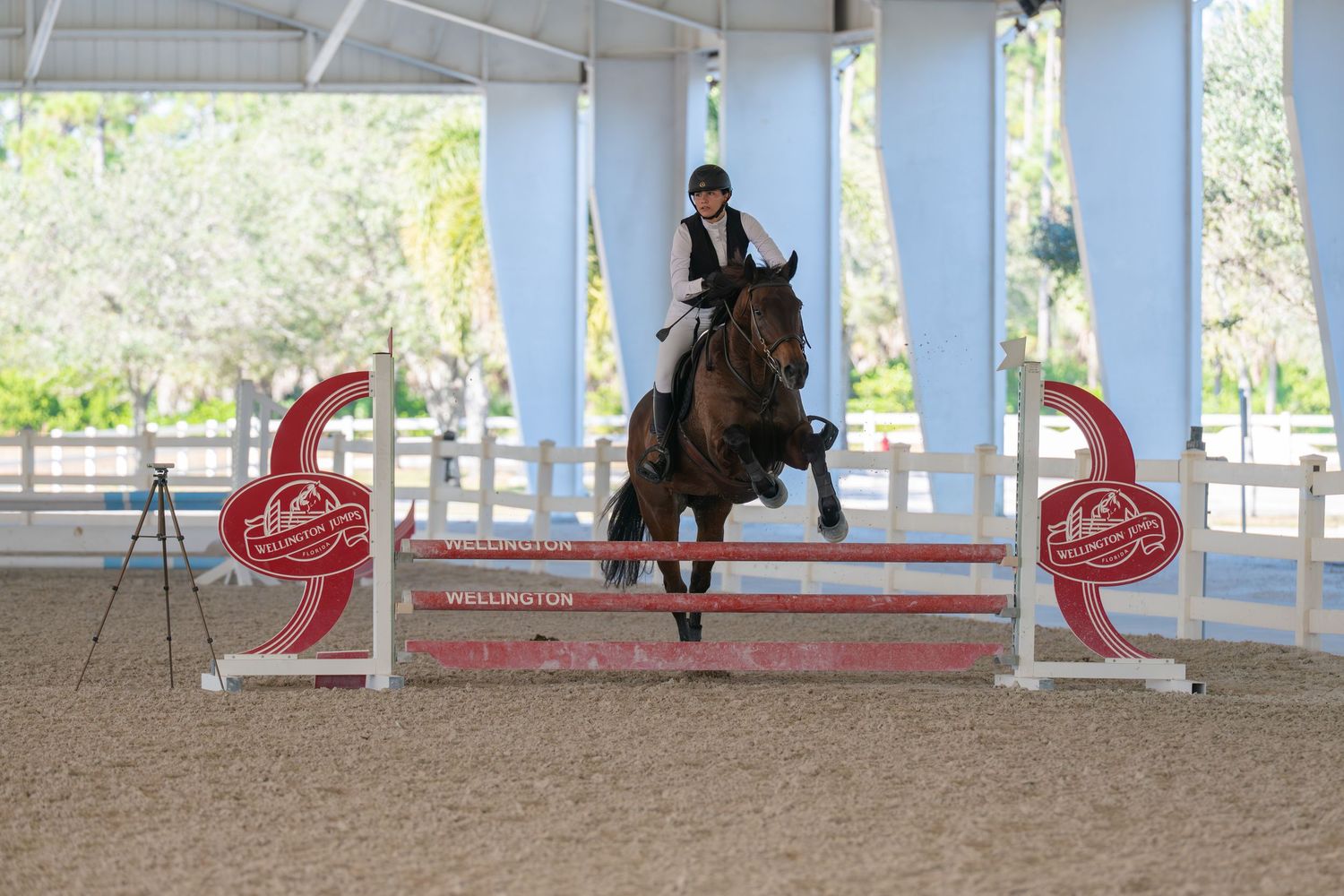 Equestrian rider jumping horse over red and white fence at Wellington indoor arena competition.