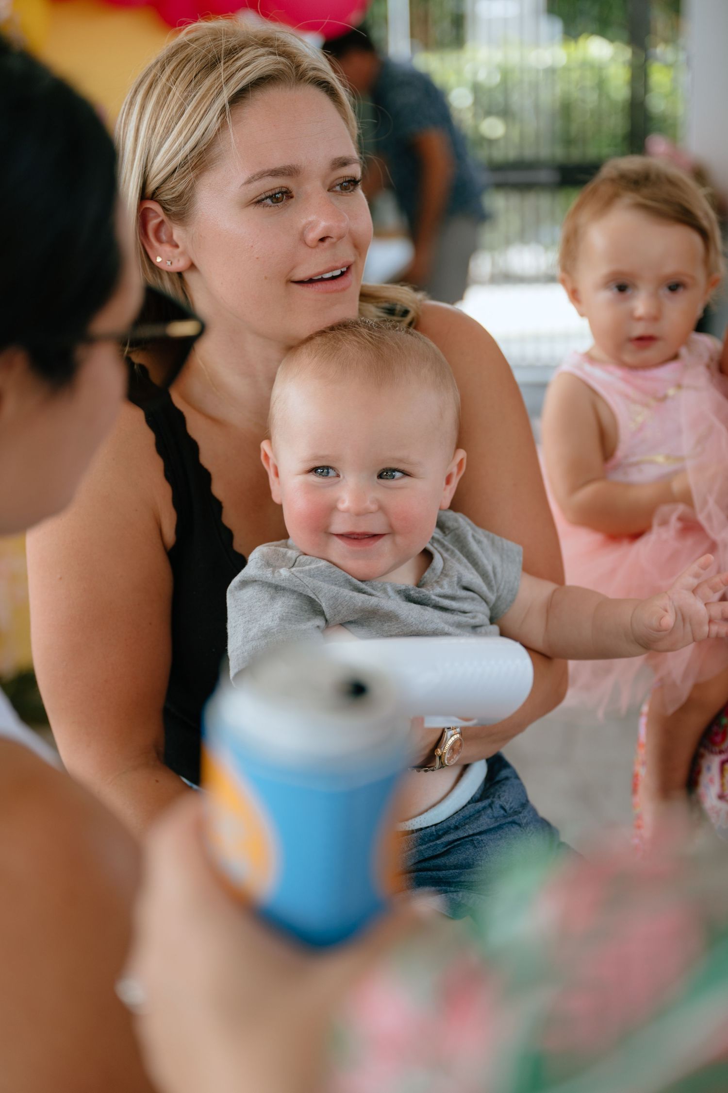 Blonde woman holding a smiling baby boy at an outdoor birthday party with a child in pink tutu nearby.