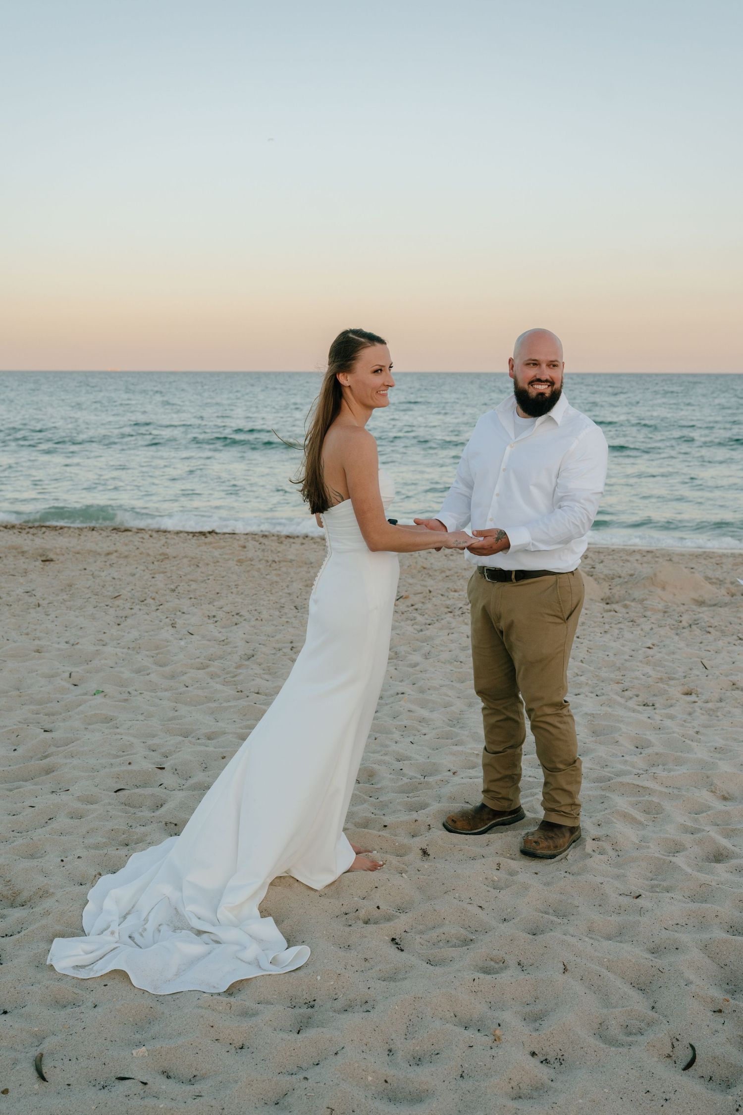 Bride and groom holding hands on a sandy beach at sunset with ocean in the background.