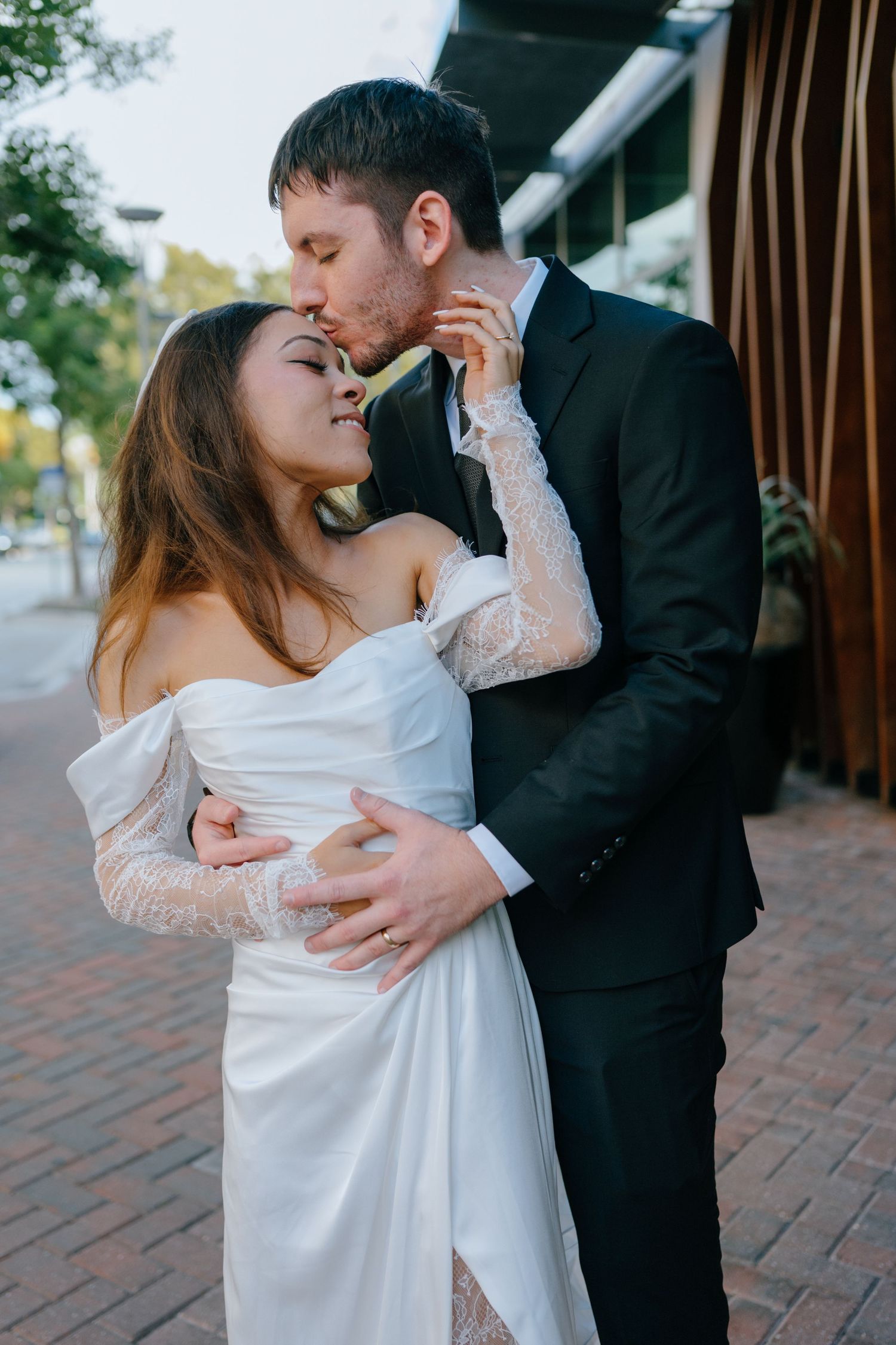 Groom in black suit kissing bride in white lace off-shoulder wedding dress outdoors.