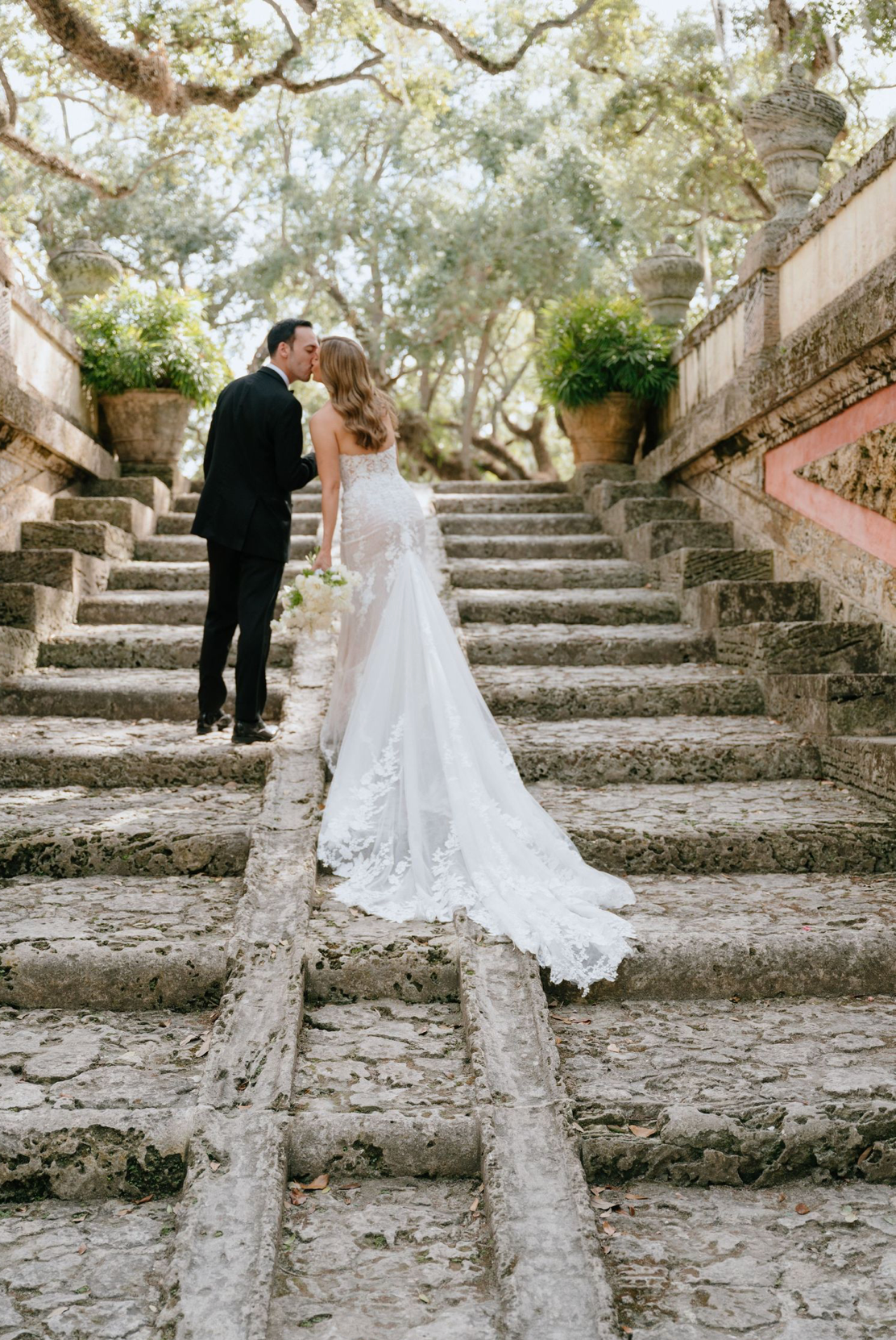 Bride and groom sharing a romantic kiss on stone steps surrounded by lush greenery at a garden wedding venue.