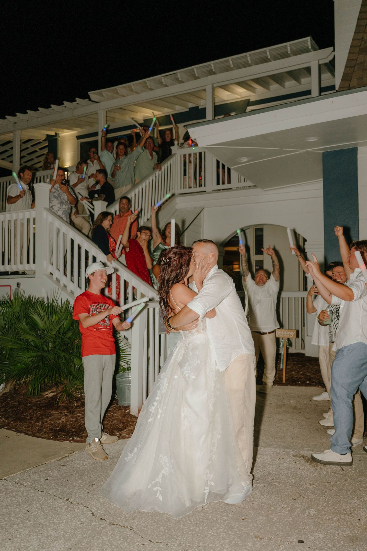 Groom lifts bride outside a white venue as cheering wedding guests line the staircase at night.