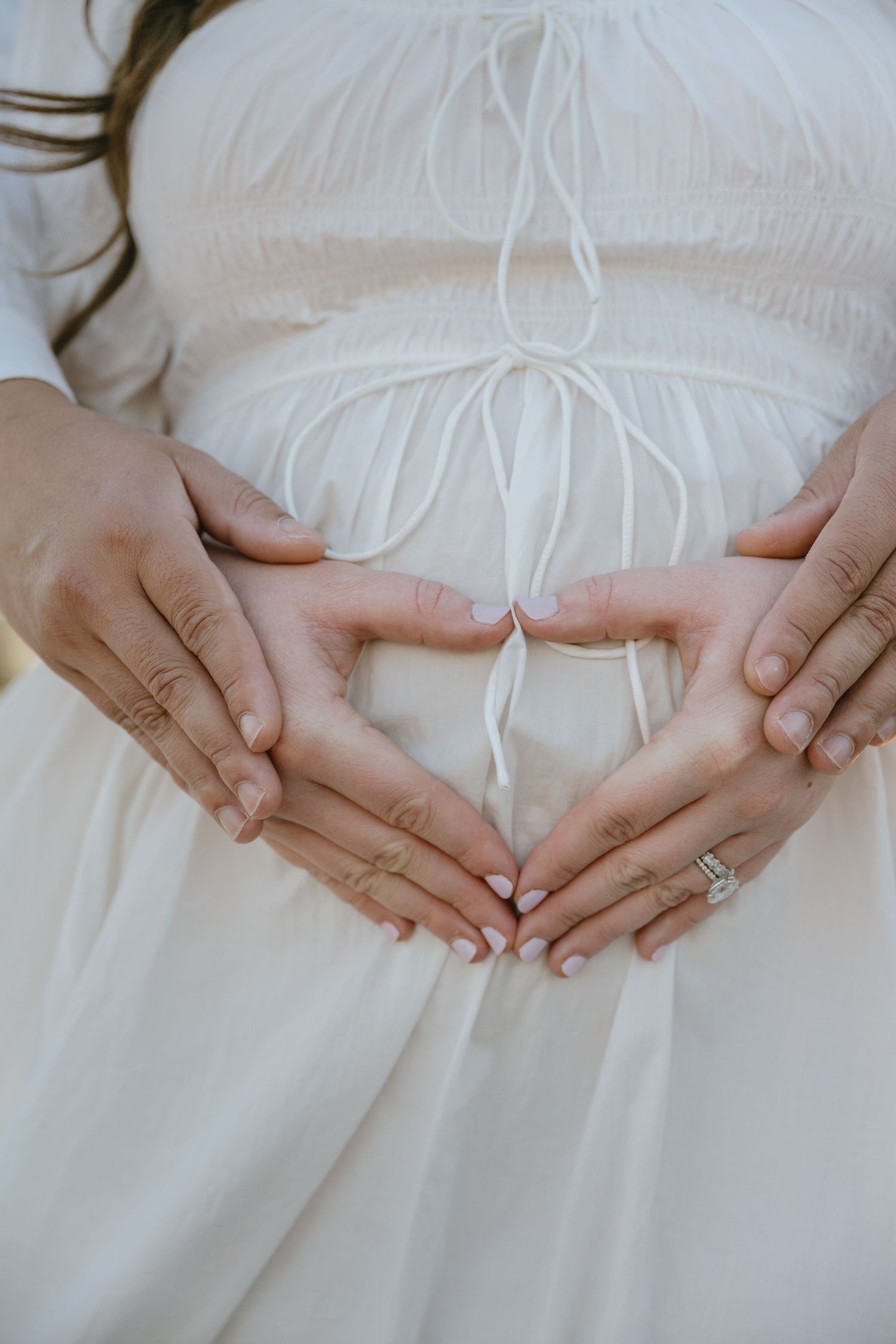 Close-up of couple forming heart shape with hands on pregnant belly in white dress.