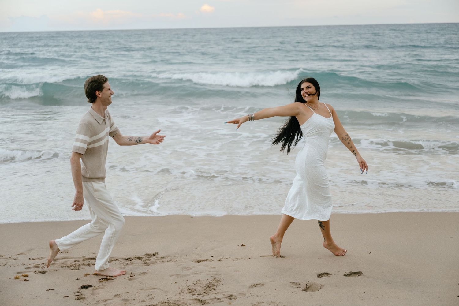 Couple playfully running on a sandy beach with ocean waves, woman in white dress and man in beige outfit.