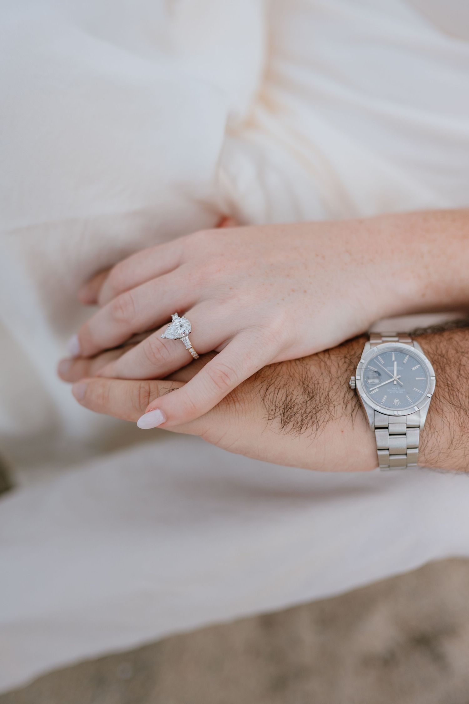 Close-up of couple's hands showing diamond engagement ring and silver watch against white fabric backdrop.