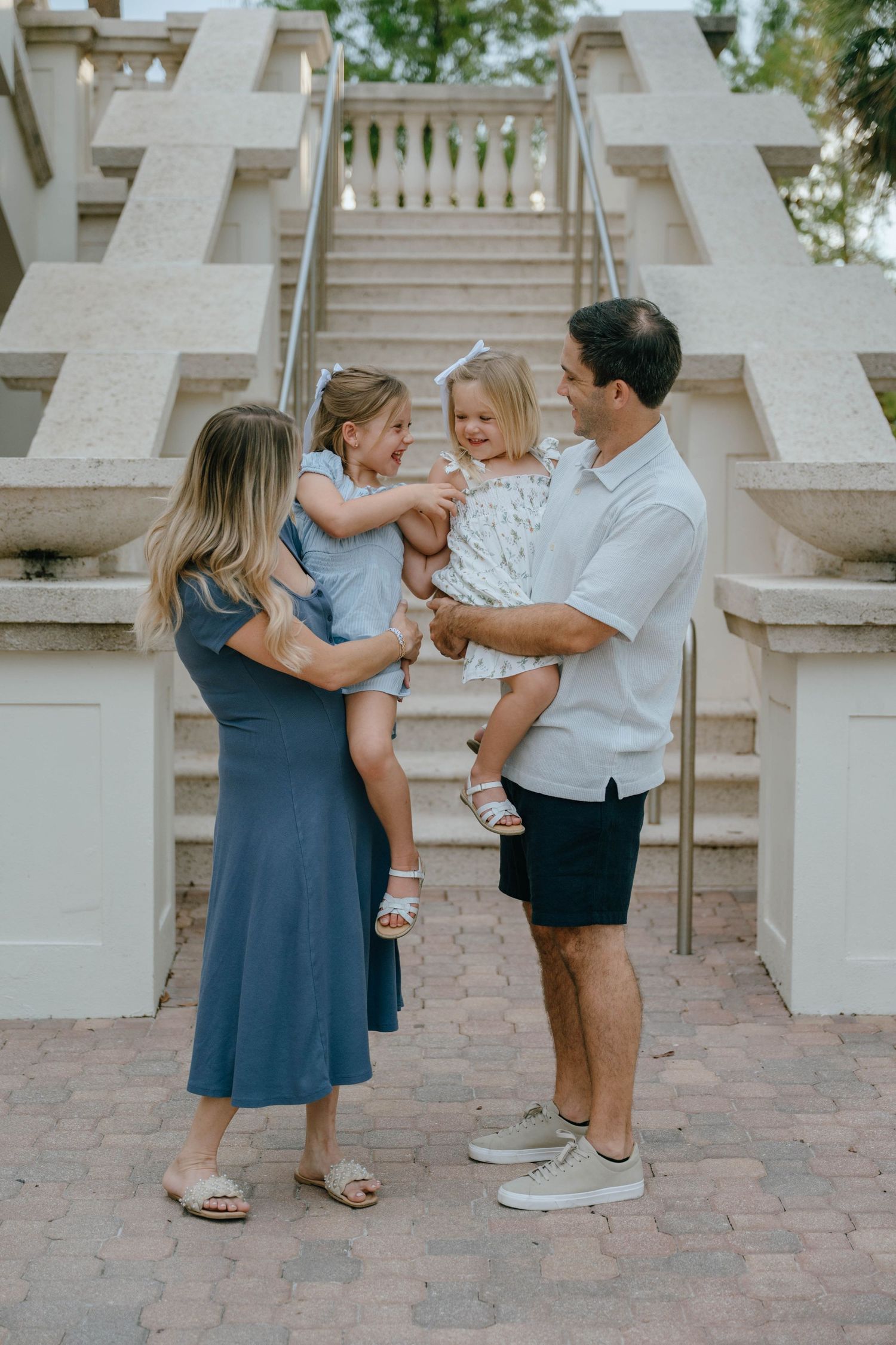 Two parents each hold one of their young daughters while the girls reach for each other playfully.