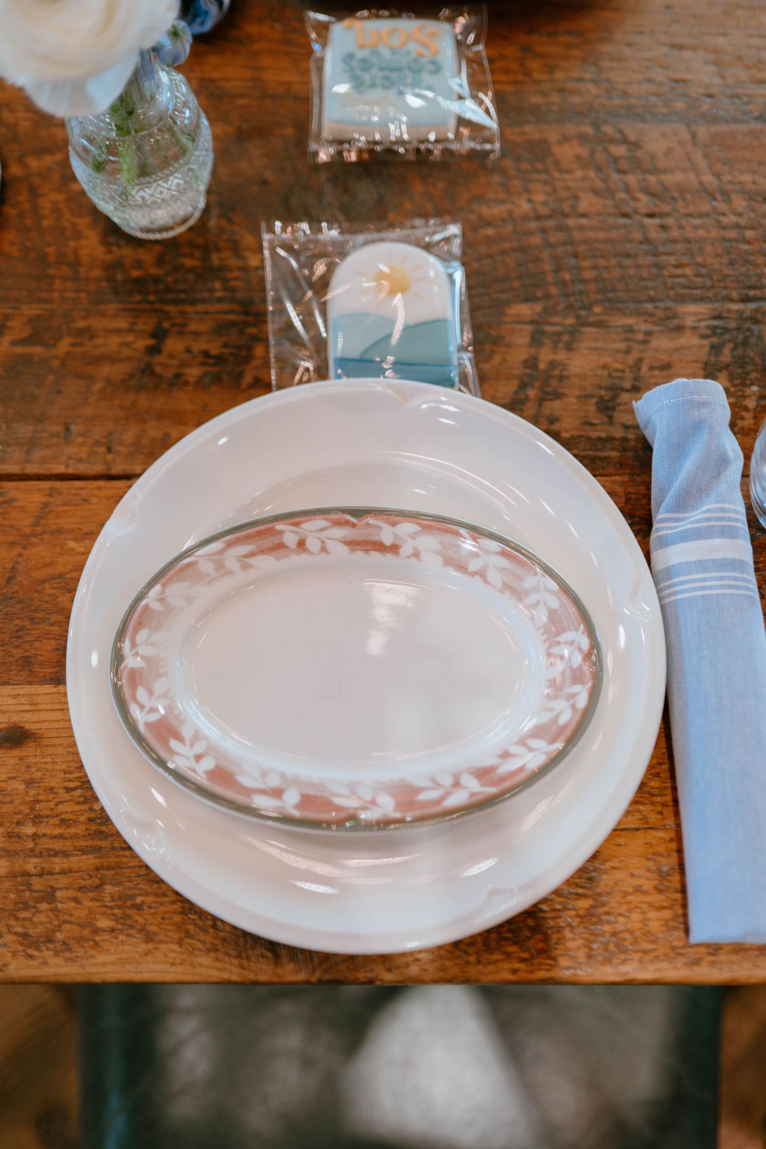 Elegant baby shower place setting with layered white plates and wrapped cookie favor on rustic wood table.