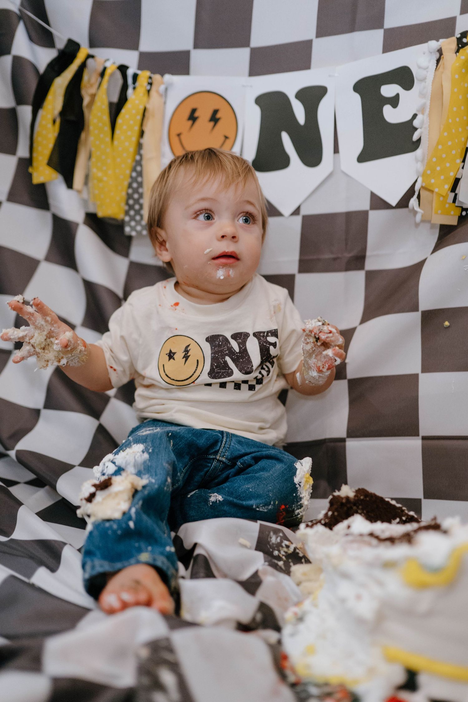 Cake-covered toddler sits on checkered backdrop with smashed birthday cake and ONE banner behind him.