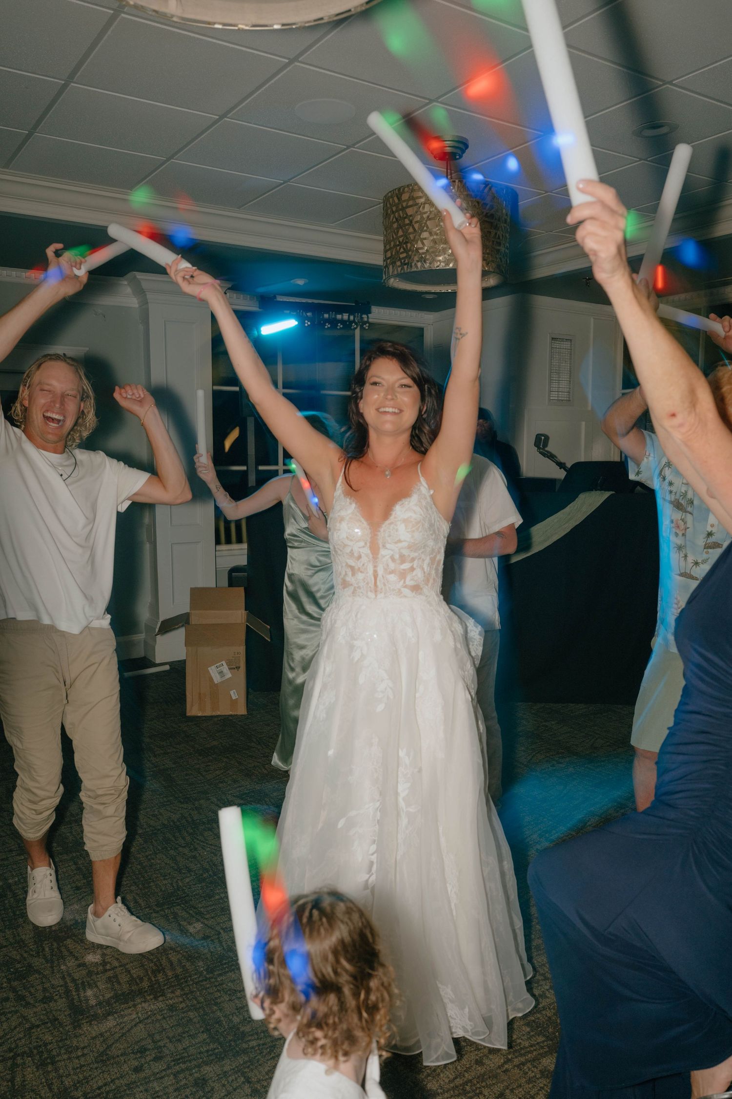 Bride in lace wedding gown celebrates with raised arms surrounded by guests holding colorful glow sticks.