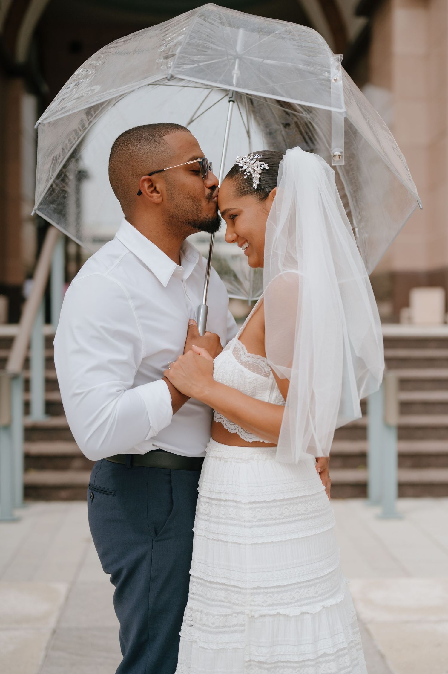 Groom kissing bride on forehead under a clear umbrella, bride wearing a lace wedding dress and veil.