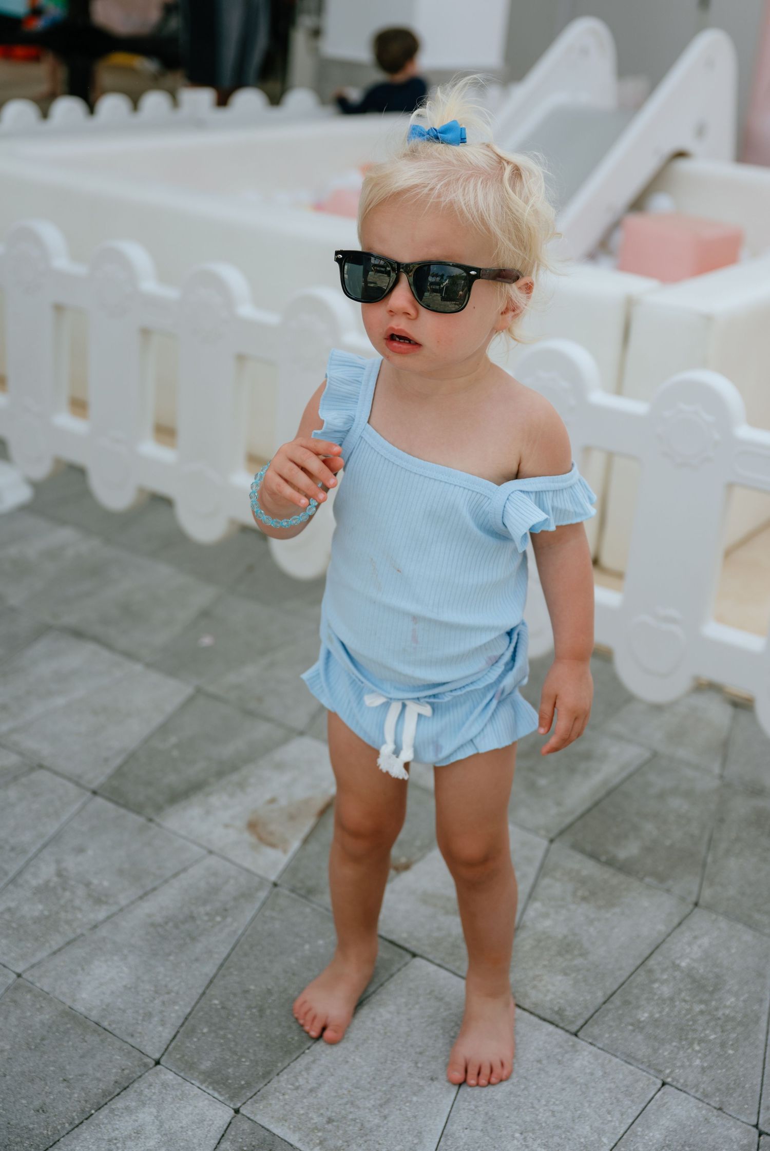 Toddler girl in light blue swimsuit and oversized black sunglasses standing barefoot on gray tile at outdoor party.