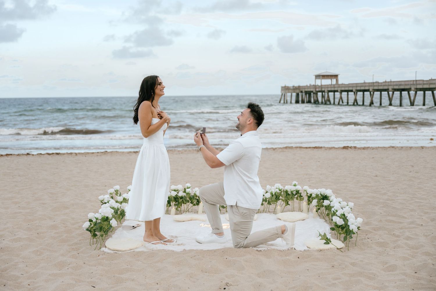 Man proposing on one knee to woman in white dress on beach with floral arrangement and pier backdrop.