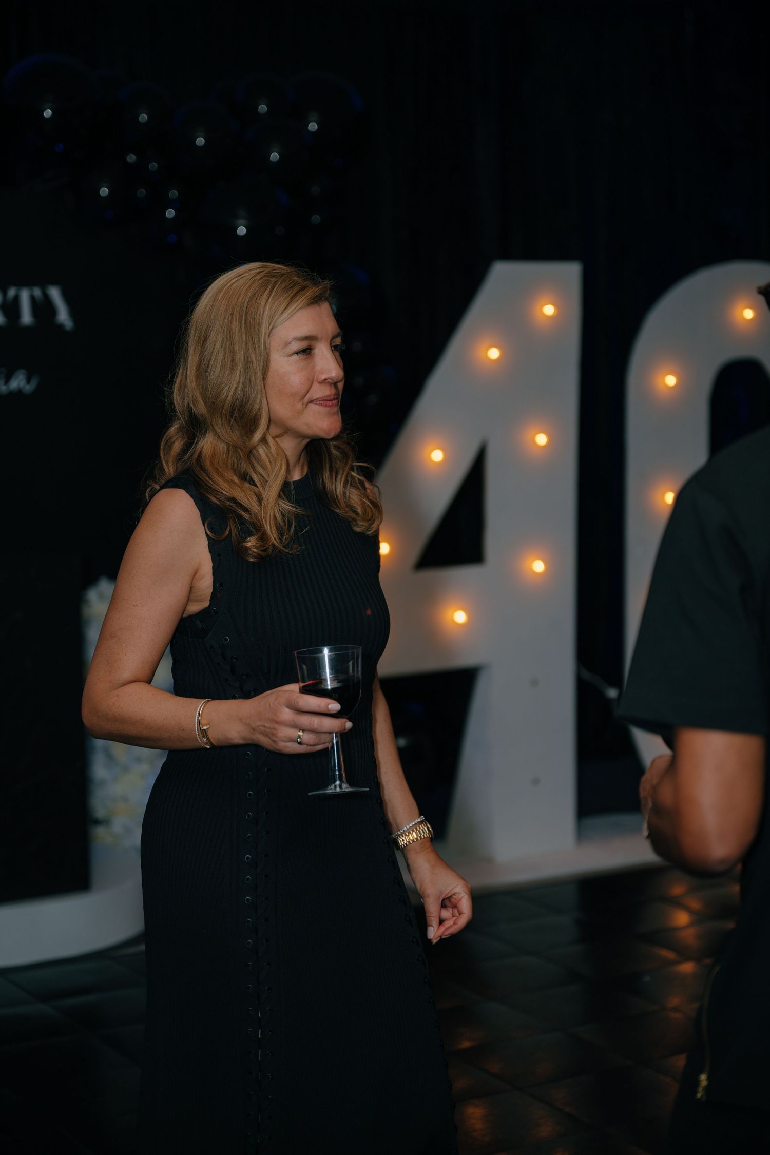 Elegant blonde woman in black dress holds a wine glass near illuminated number 40 birthday sign.