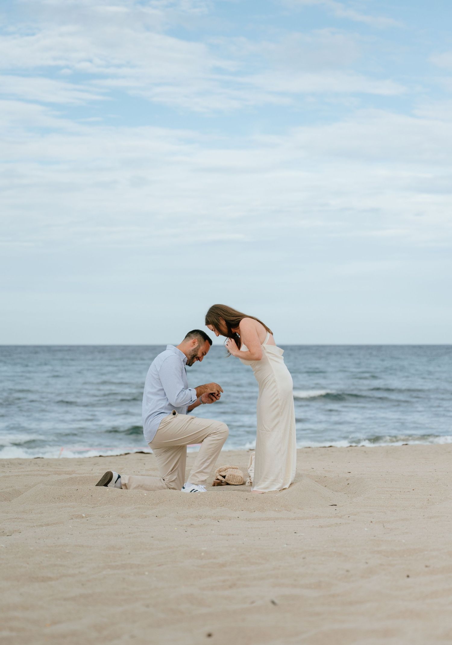 Man proposing on one knee to woman in white dress on a sandy beach with ocean backdrop.