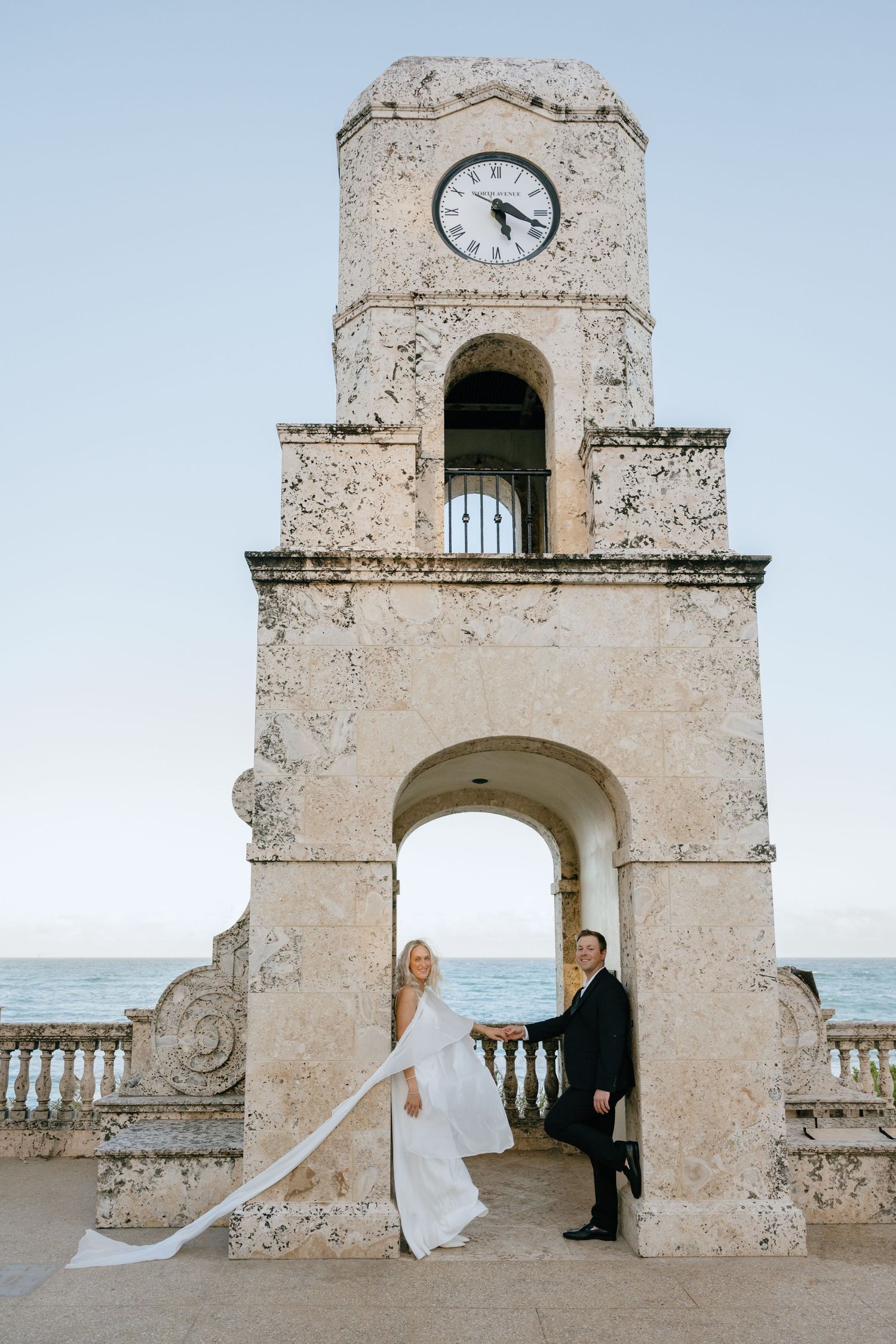Bride and groom pose beneath a historic stone clock tower with ocean views at golden hour.