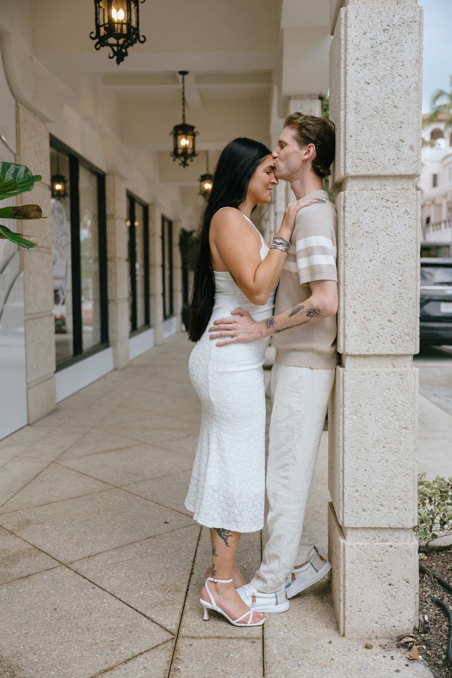 Tattooed couple sharing an intimate moment leaning against a stone column in an elegant covered walkway.