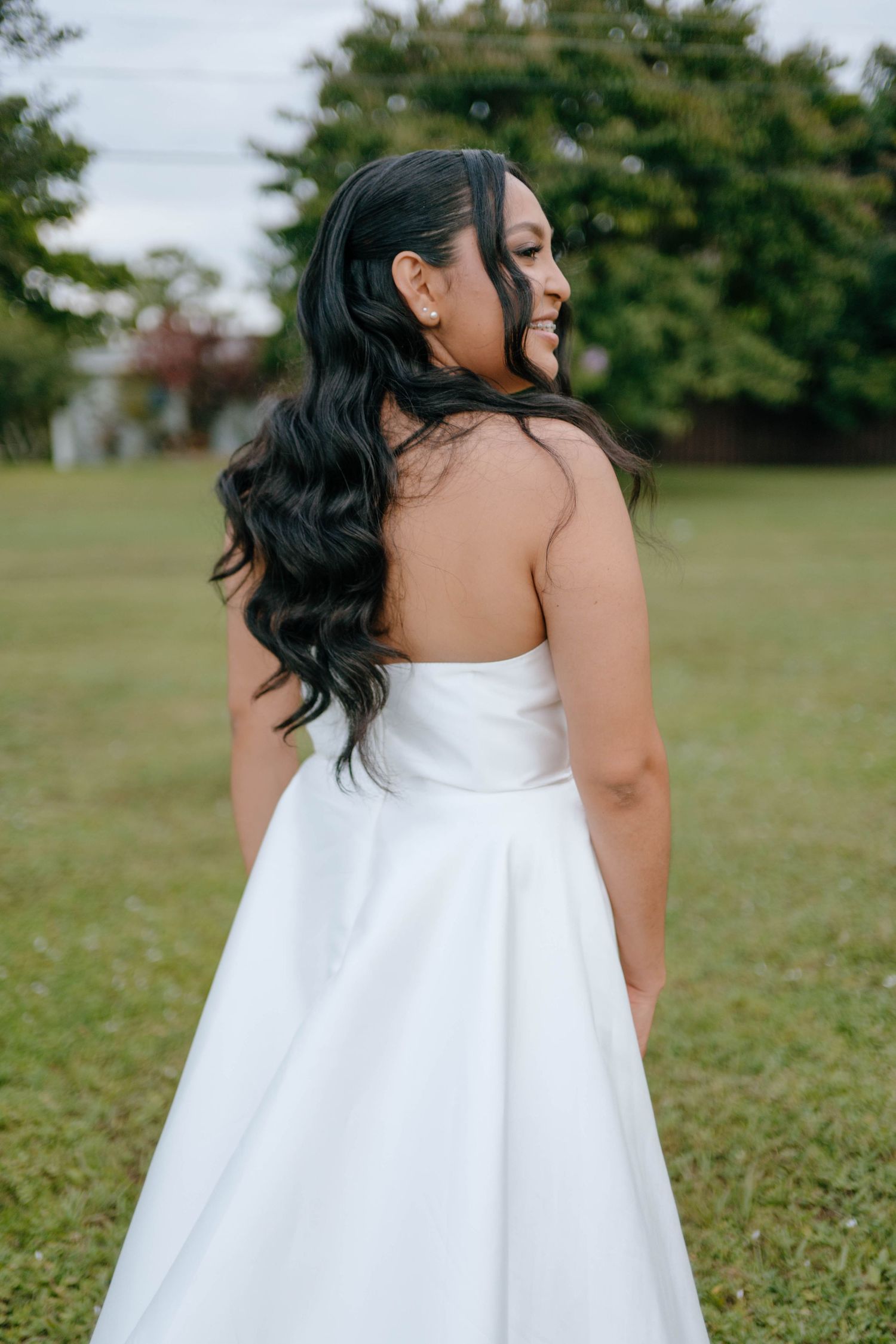 Bride with long dark wavy hair in strapless white satin wedding gown posing outdoors on green lawn.