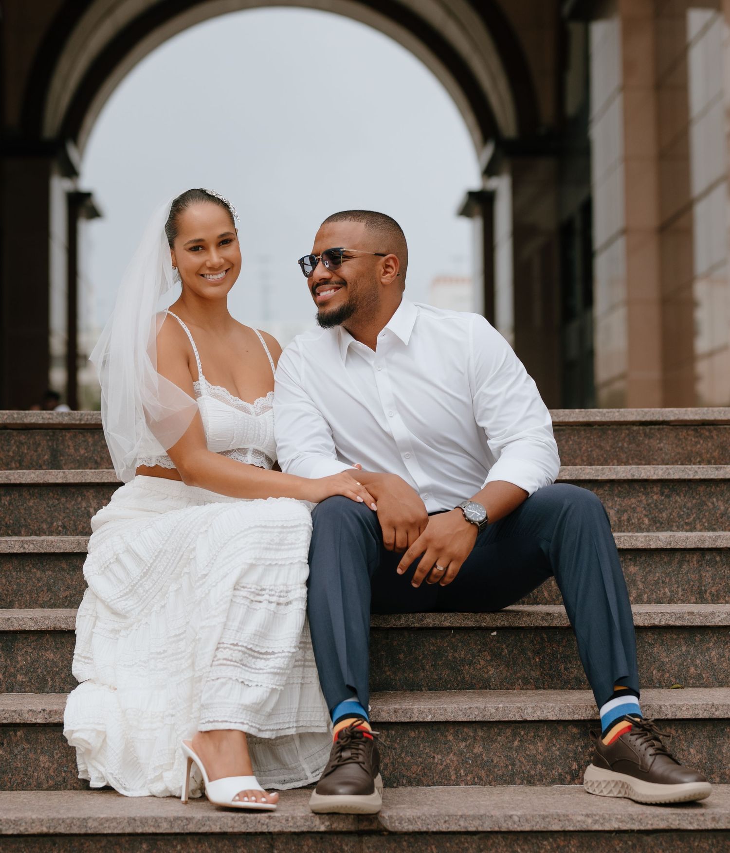 Happy newlywed couple sitting on stone steps, bride in white dress with veil and groom in white shirt.