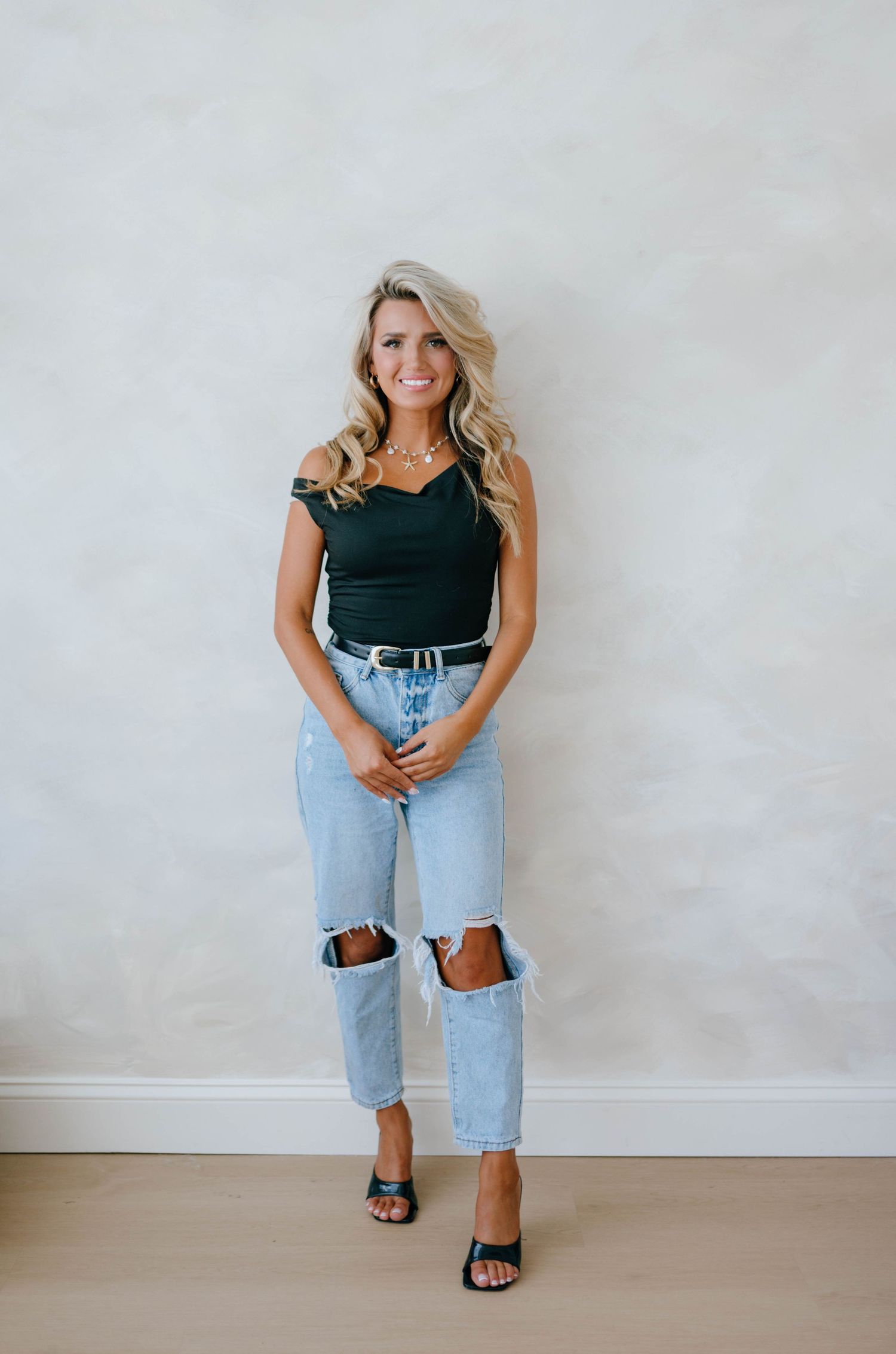 Blonde woman in black off-shoulder top and ripped jeans standing against white wall.