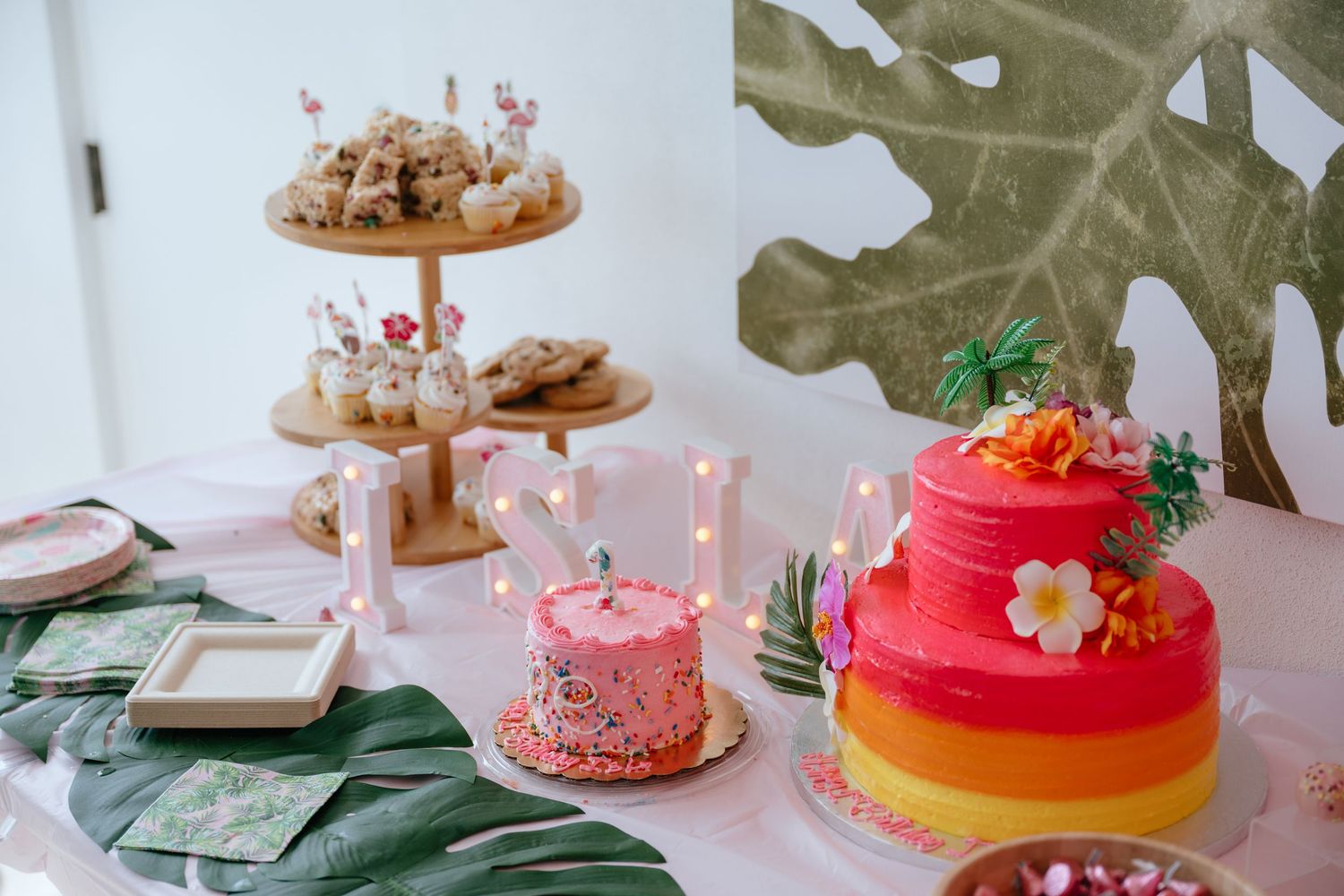 Colorful tropical-themed first birthday party table with tiered cake, pink smash cake, and festive decorations.