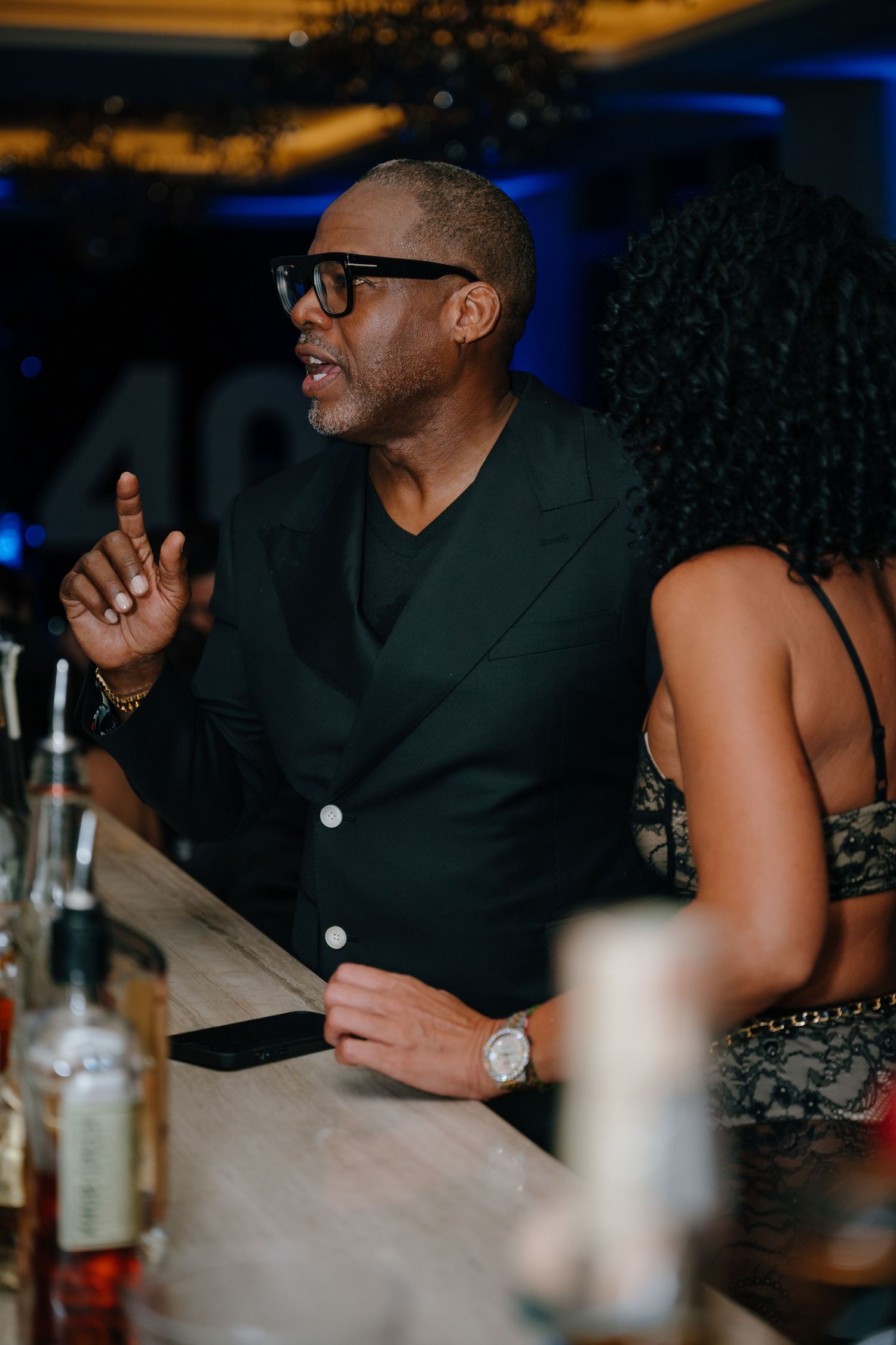Stylish man in black suit and sunglasses gestures while talking at a bar during an evening event.