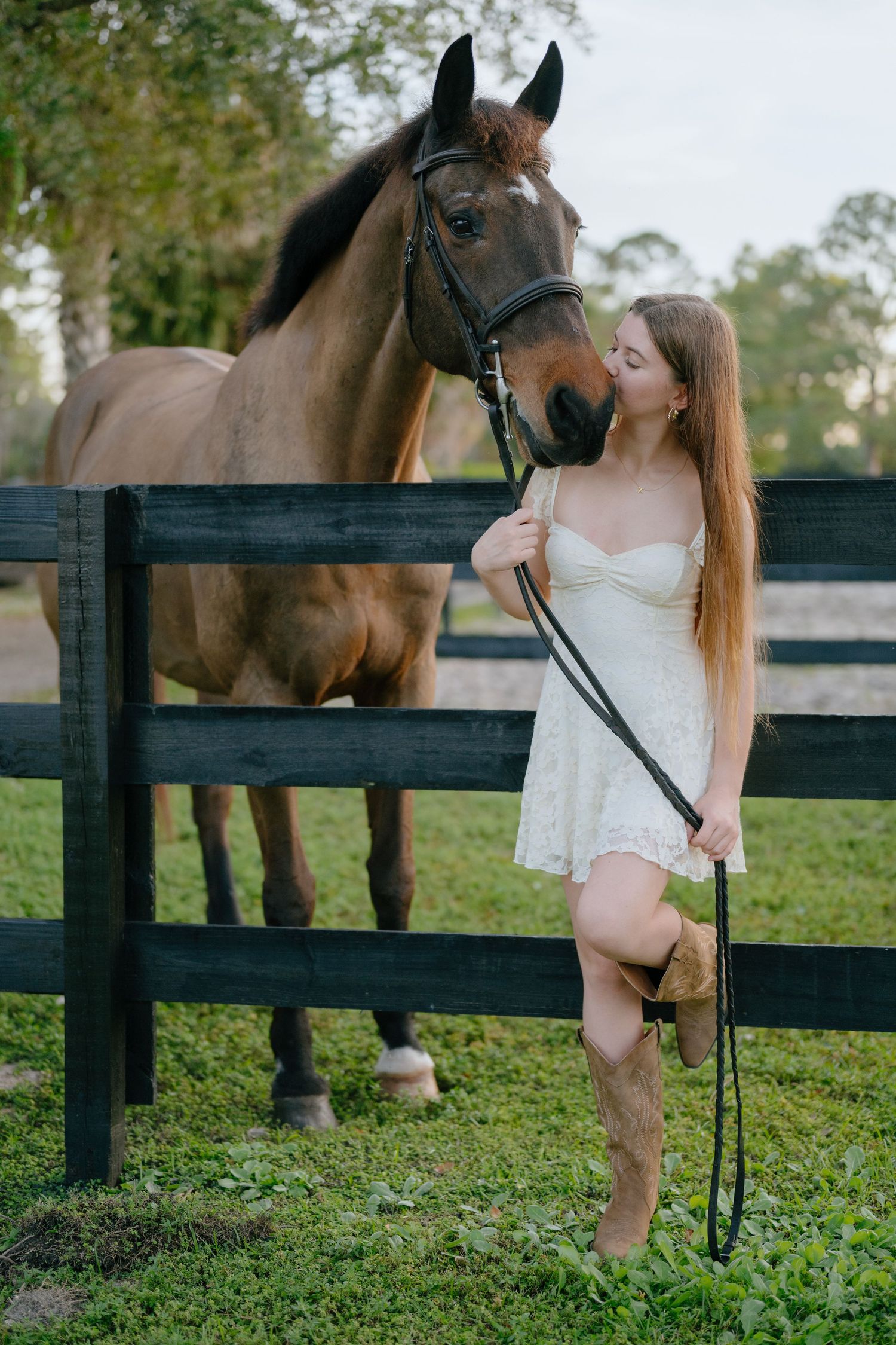 Young woman in white lace dress kisses a bay horse standing by a dark wooden farm fence.