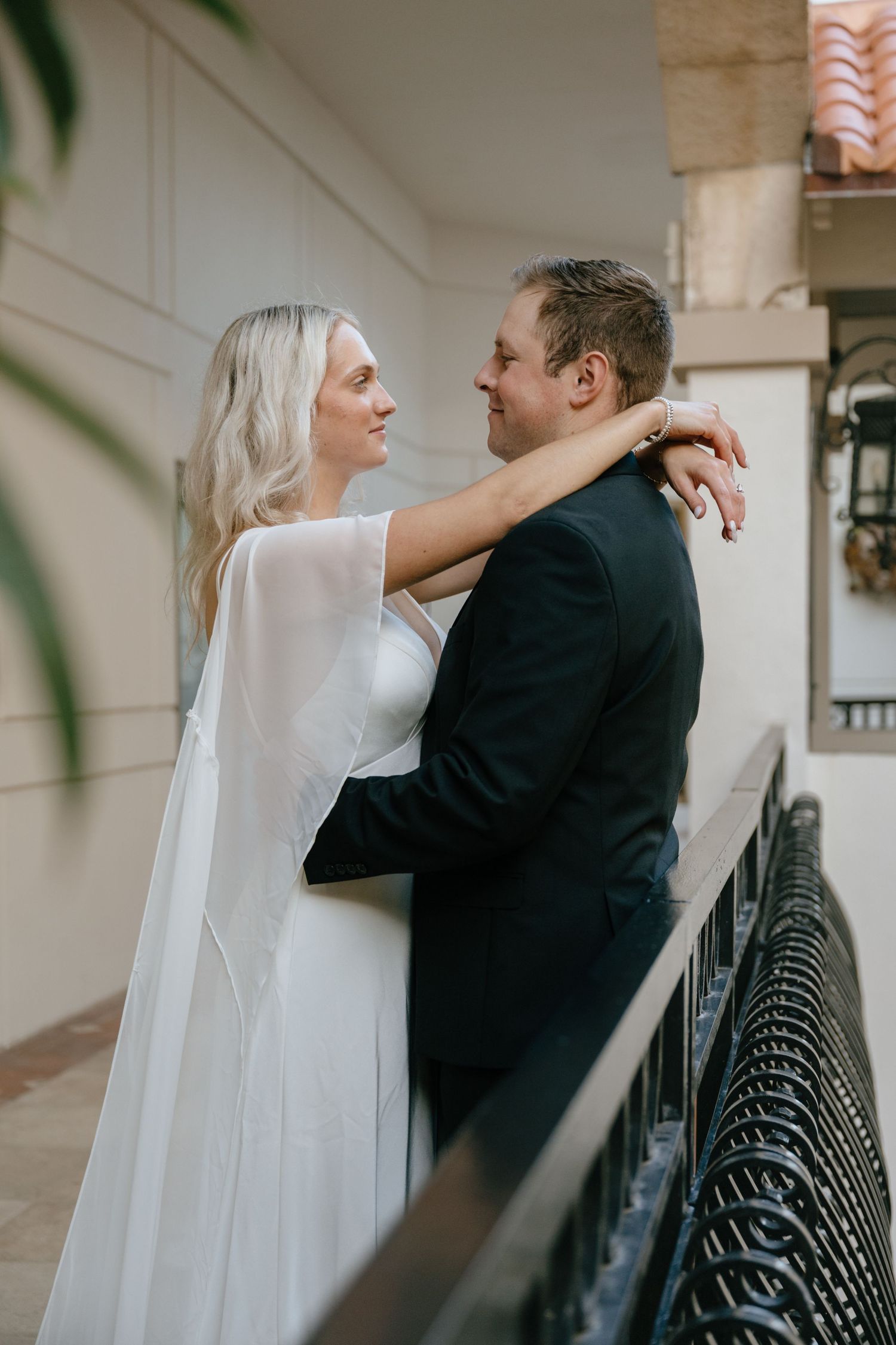 Bride and groom share a tender embrace on a balcony, bride in white cape gown, groom in black suit.