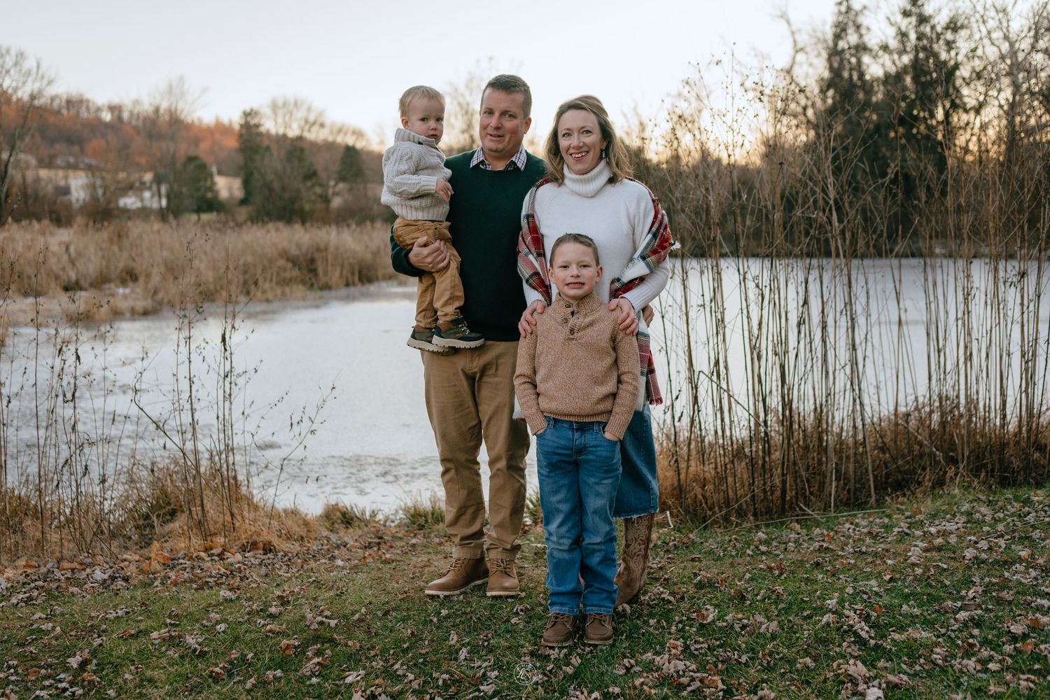 Family of four smiling together near a peaceful pond surrounded by autumn foliage at golden hour.