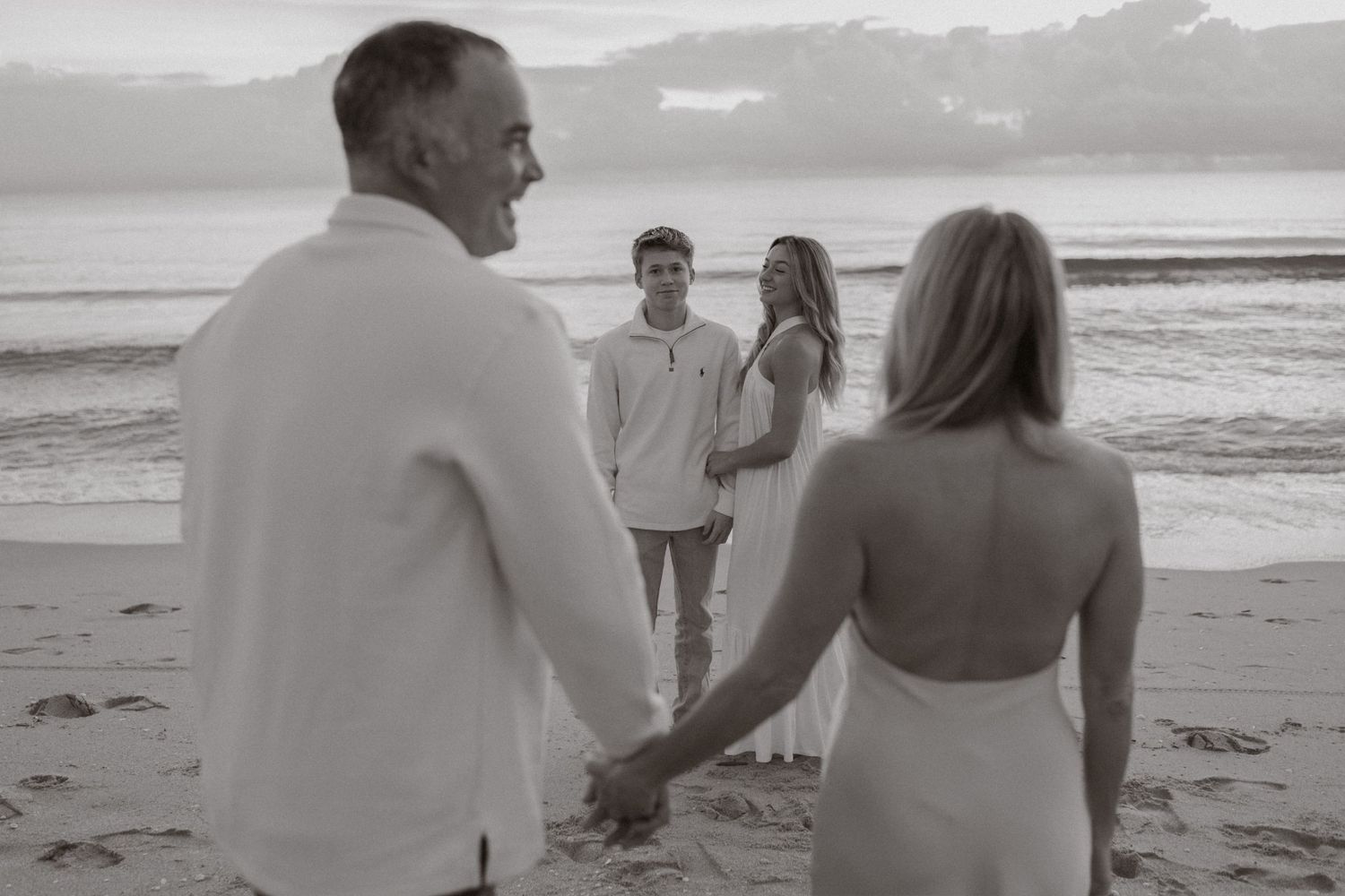Black and white photo of parents holding hands facing their teens on a beach.