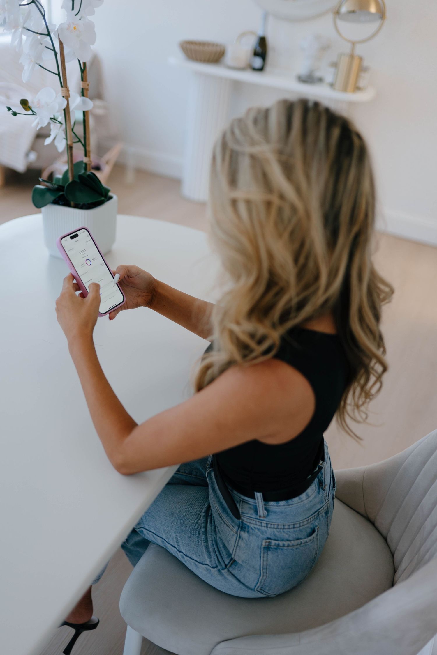 Blonde woman in black top and jeans using a smartphone while seated at a white table with orchids nearby.