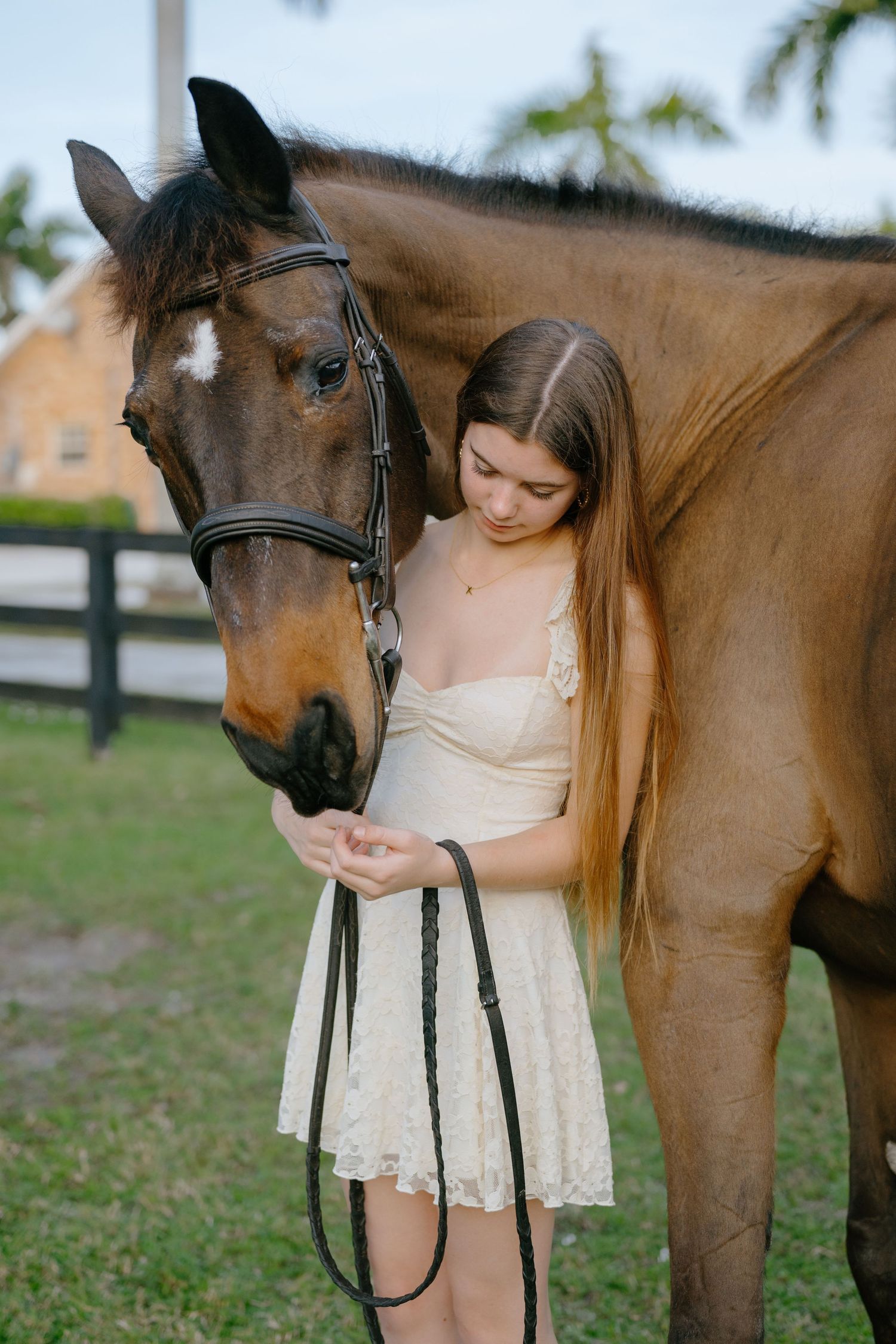 A young woman in a cream lace dress gently cradles the face of a bay horse in an outdoor setting.