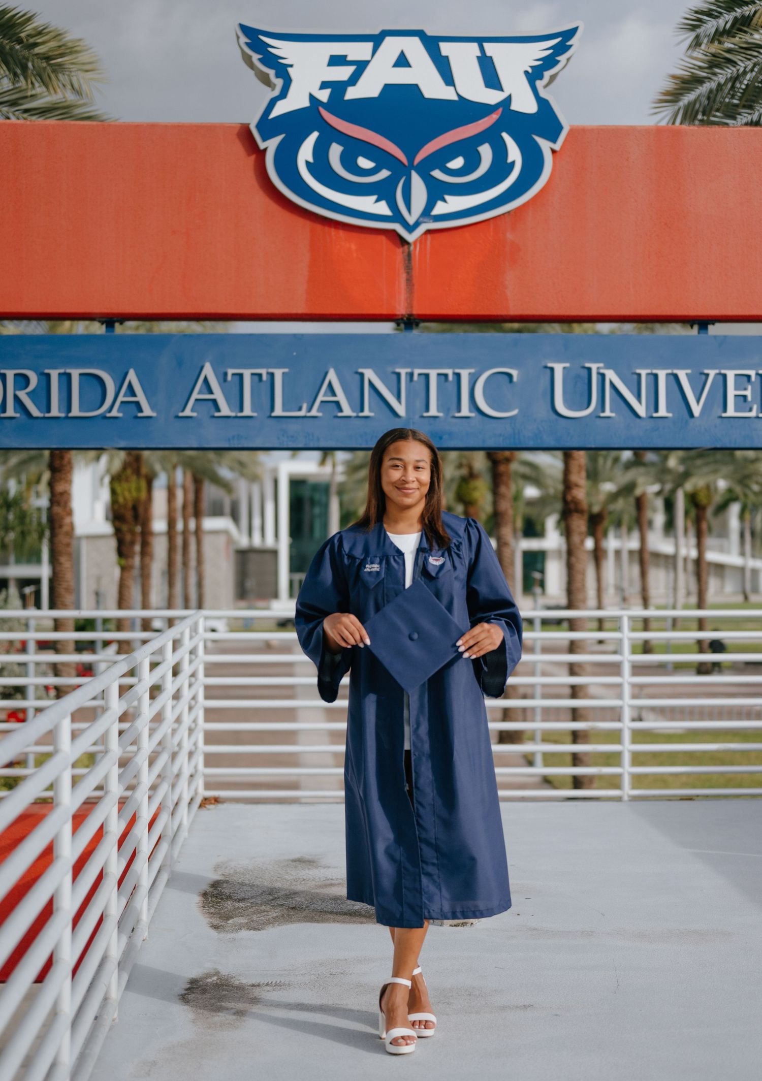 Graduate in navy FAU cap and gown posing under Florida Atlantic University owl logo sign.