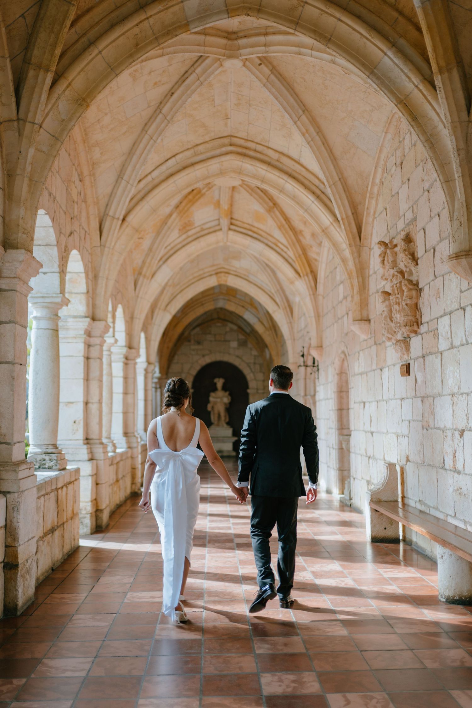 Bride and groom walking hand in hand through a stunning Gothic stone corridor.