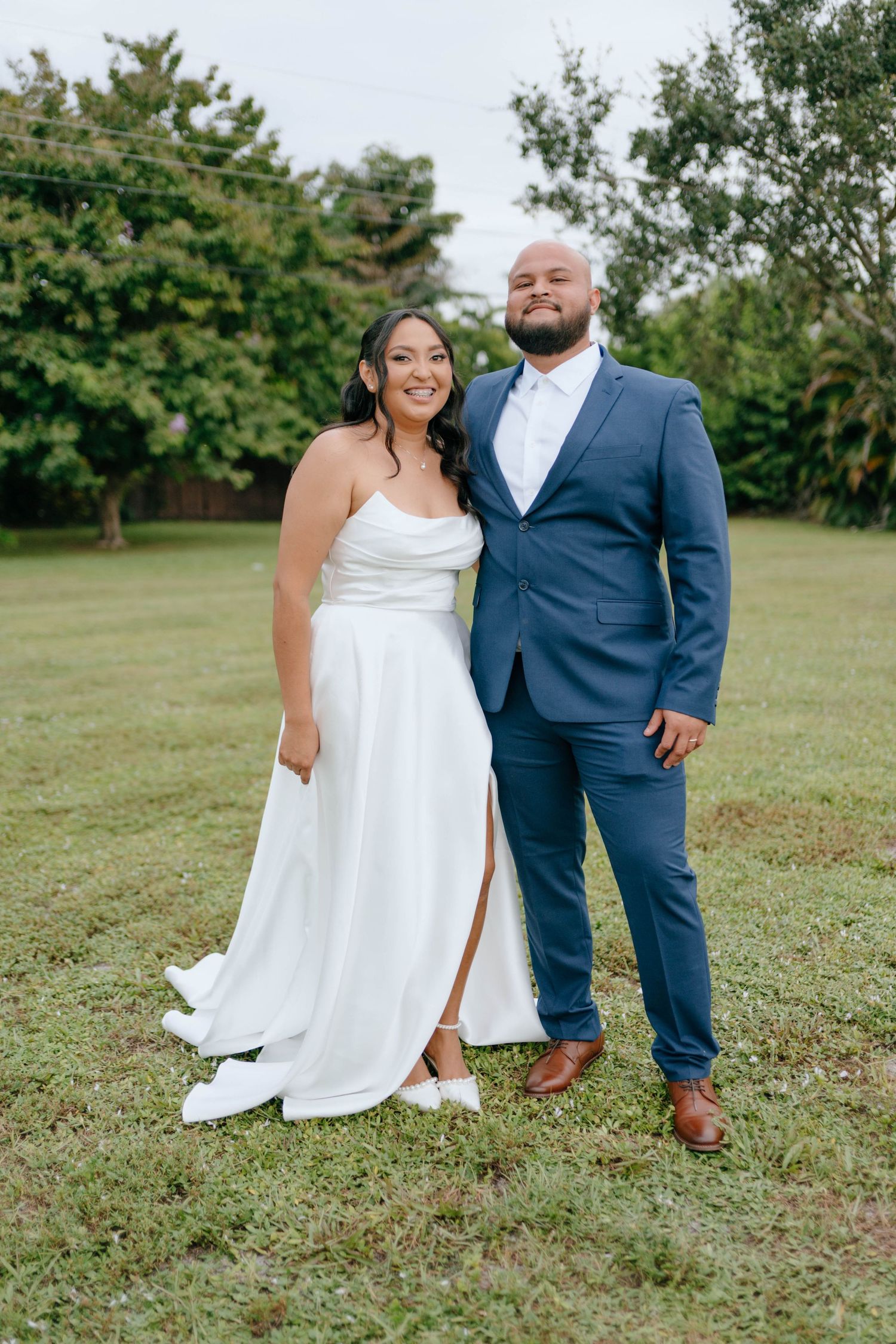 Bride and groom pose together on lush green lawn, groom wearing navy blue suit on wedding day.