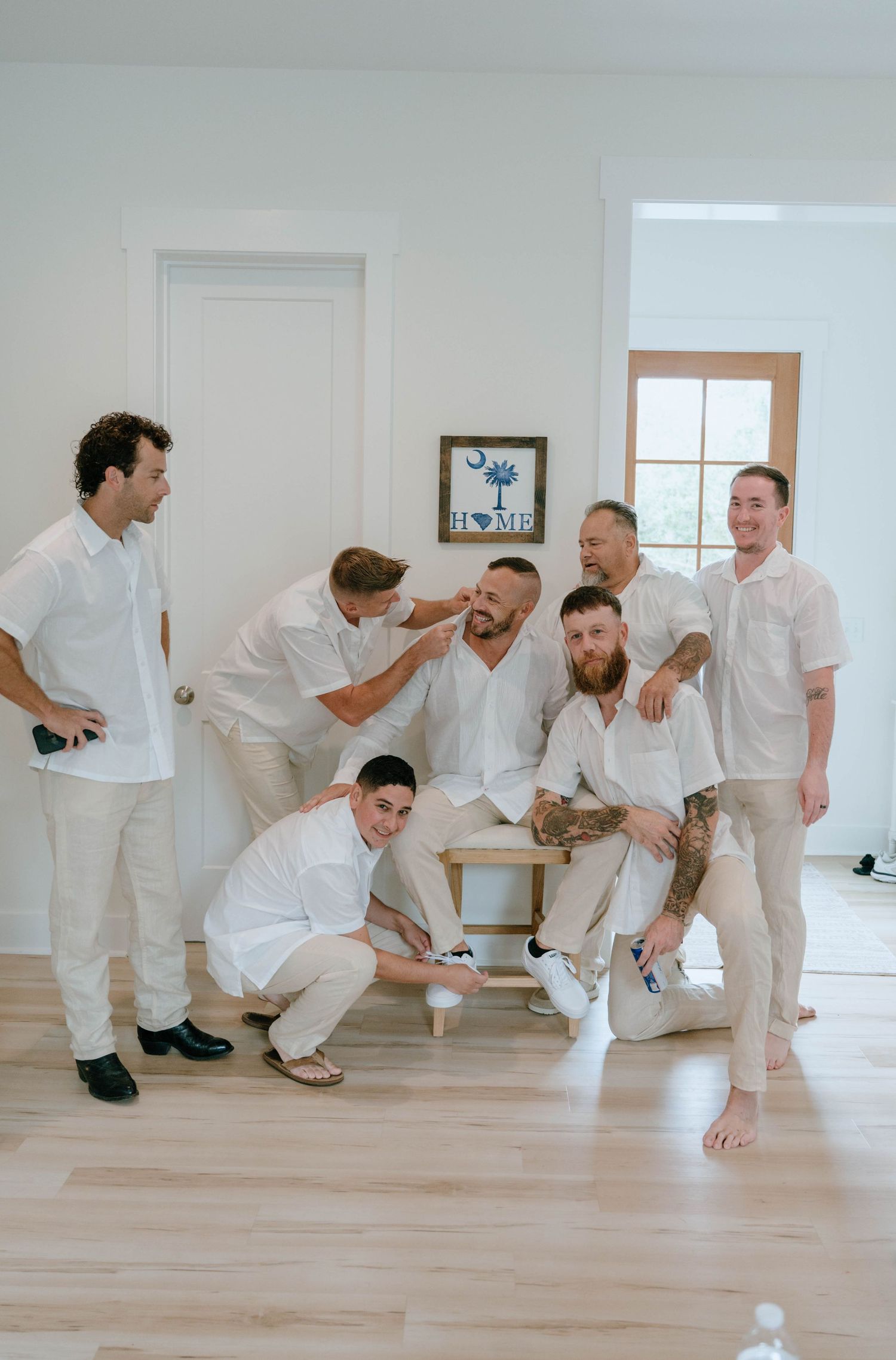 Groom and groomsmen laughing together in matching white shirts and khaki pants indoors.