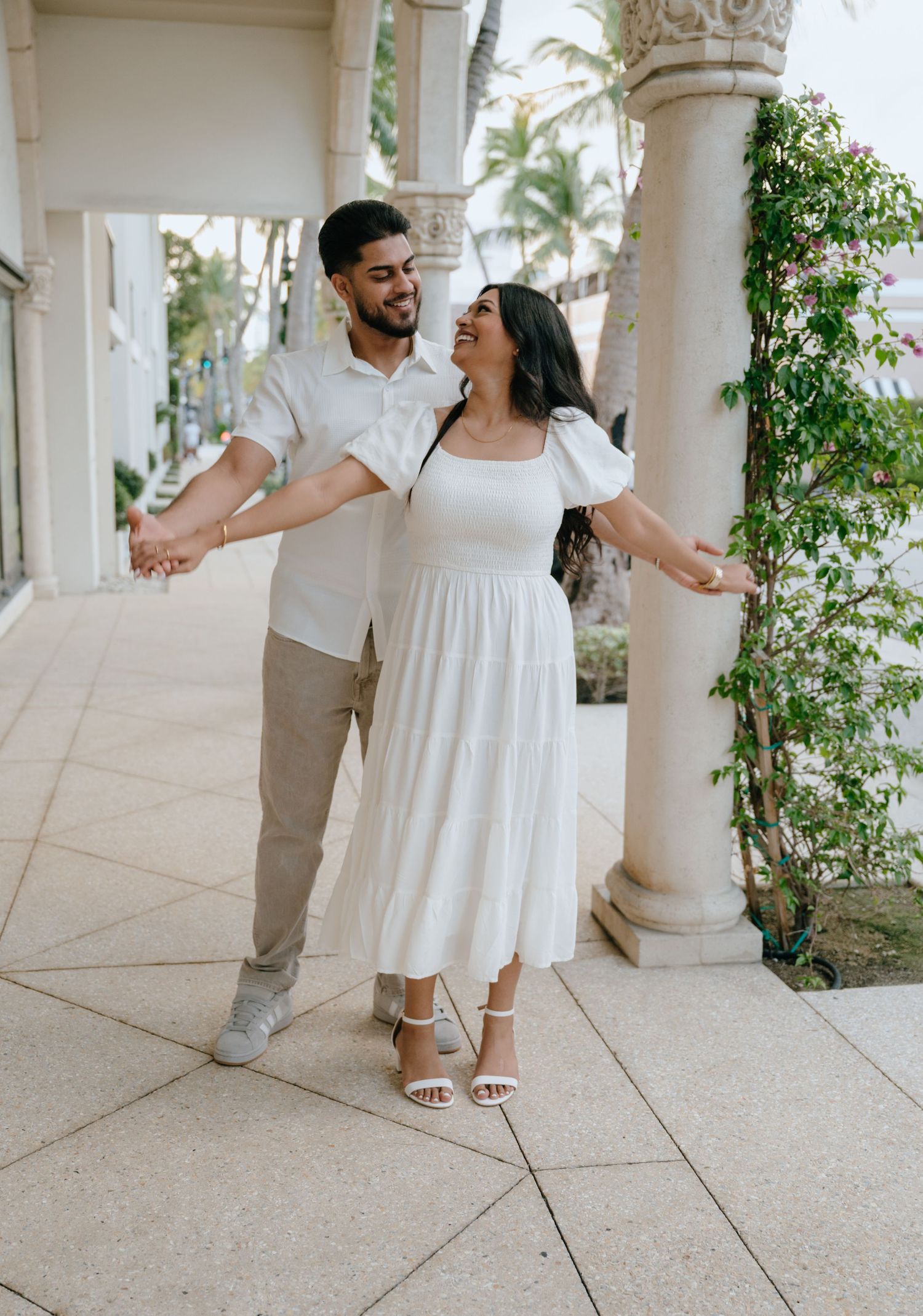 Joyful couple in white outfits dancing and laughing in a sunlit outdoor colonnade.