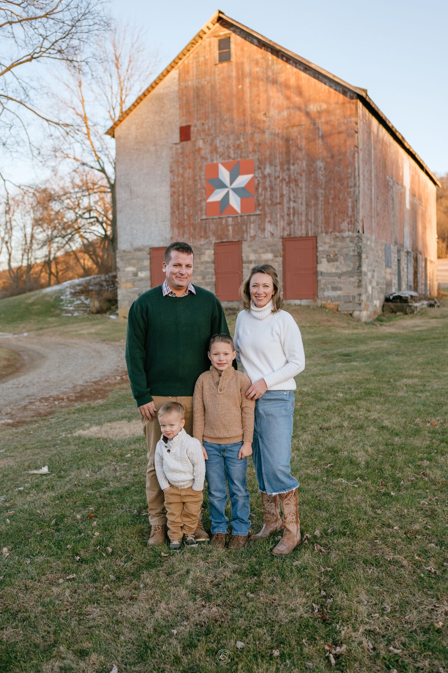 Family of four posing in front of a rustic red barn with a quilt square decoration during golden hour.