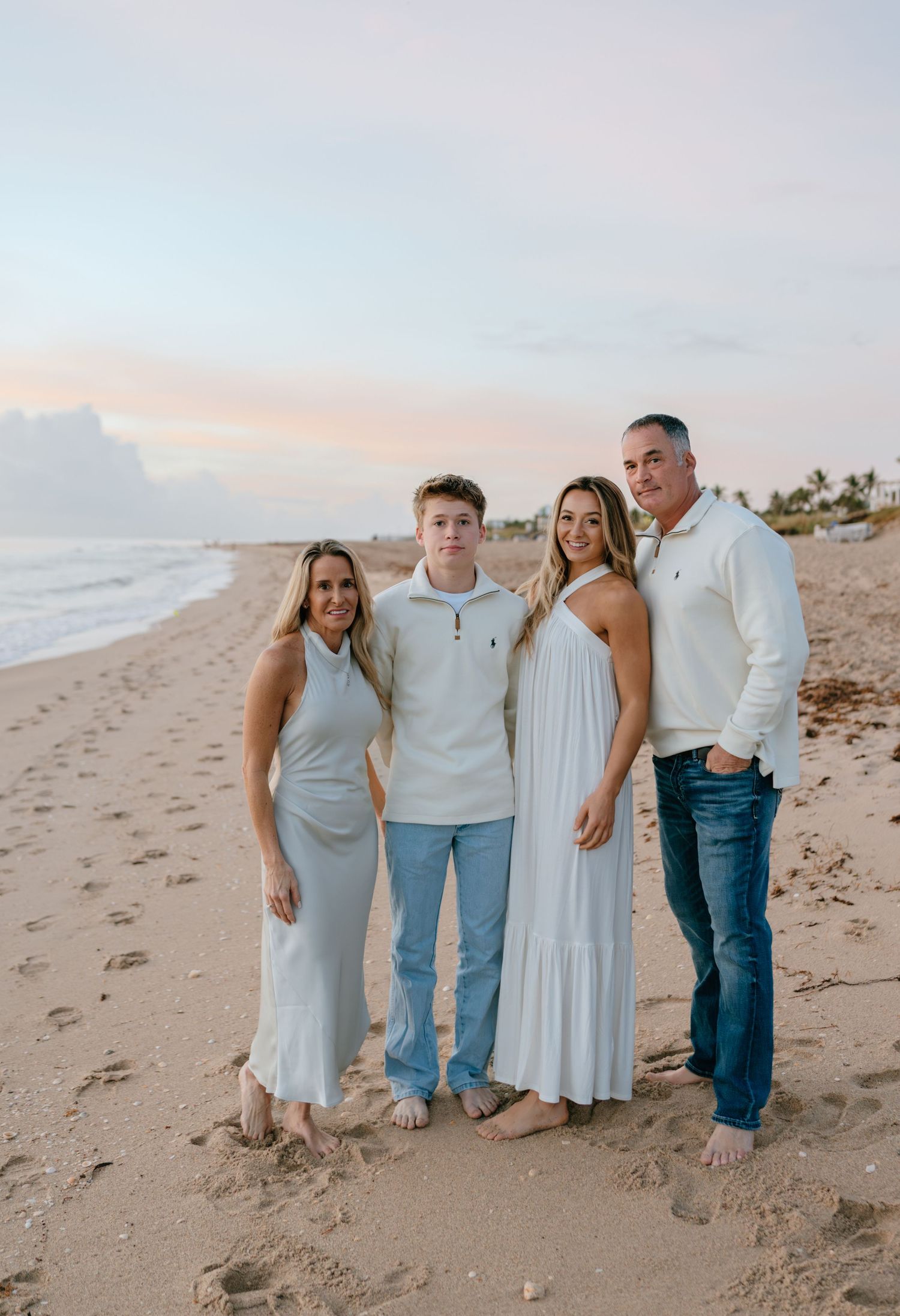 A family of four poses together smiling on a beach at sunset.